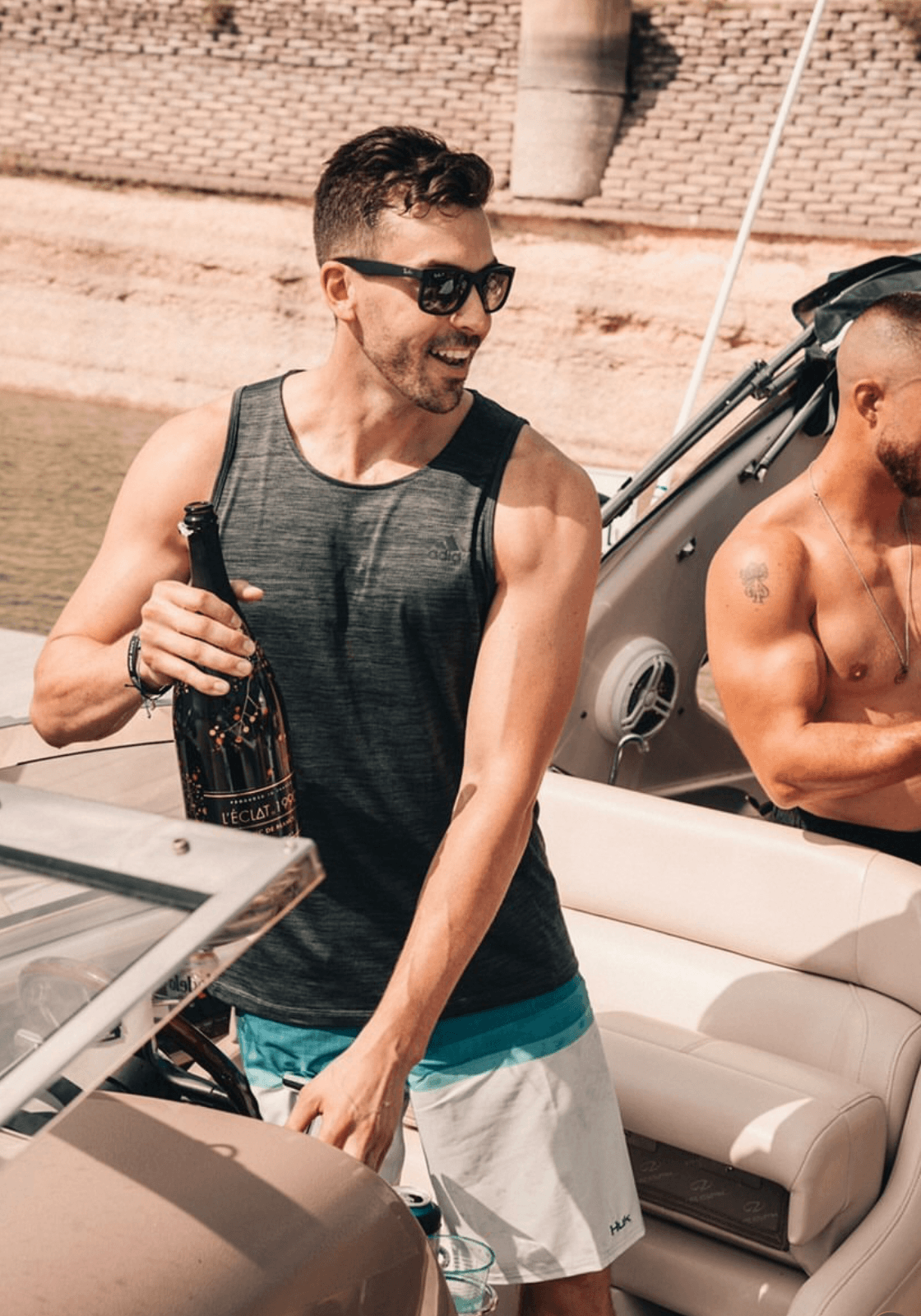 Man wearing sunglasses and a sleeveless shirt holding a bottle on a boat, smiling, with another shirtless man nearby.