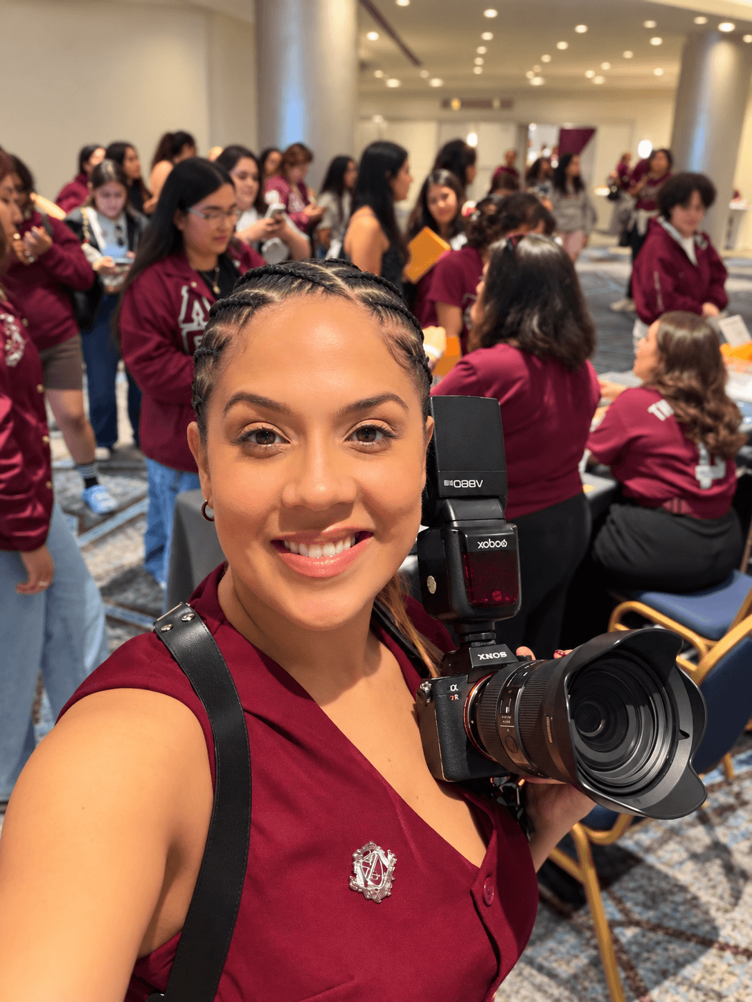 Young woman smiling with camera, group of people in maroon uniforms in background, indoor setting.