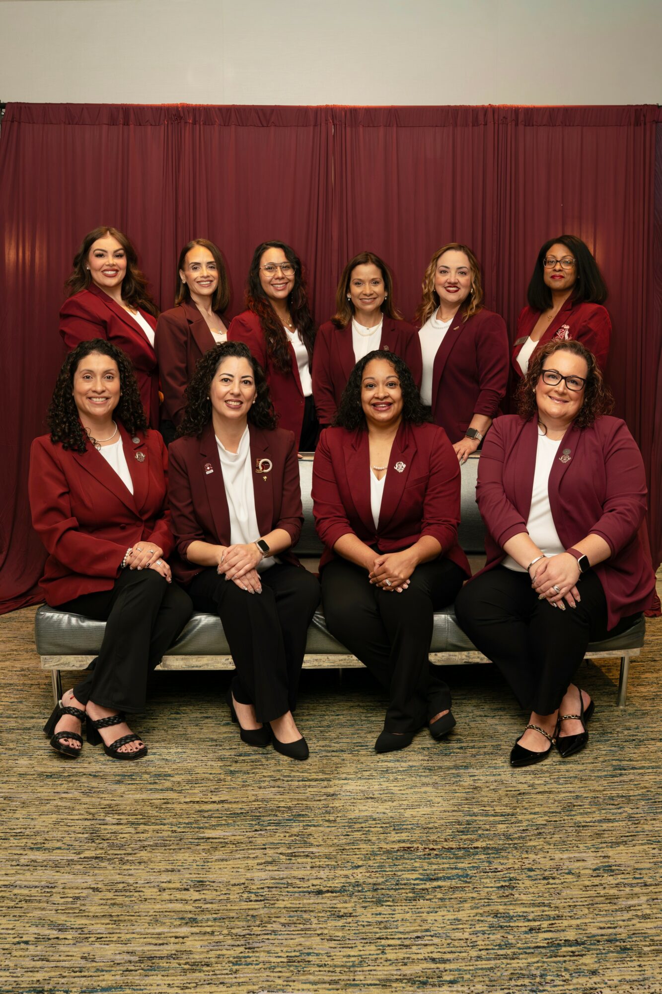 Ten women in matching maroon blazers and black pants pose in front of a red curtain, smiling for a group photo.