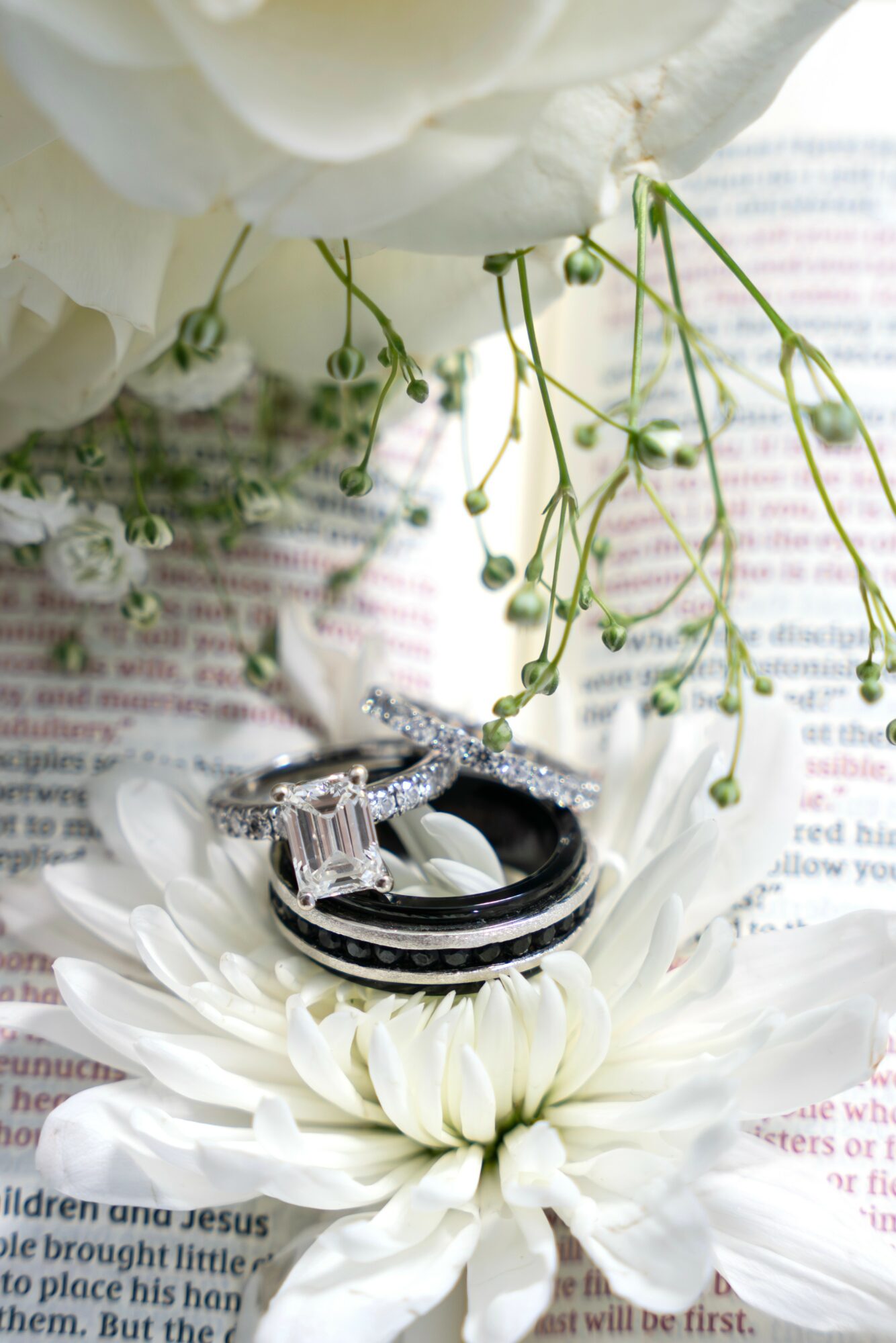 Wedding rings on a white flower with green stems and small white flowers, placed on printed paper.