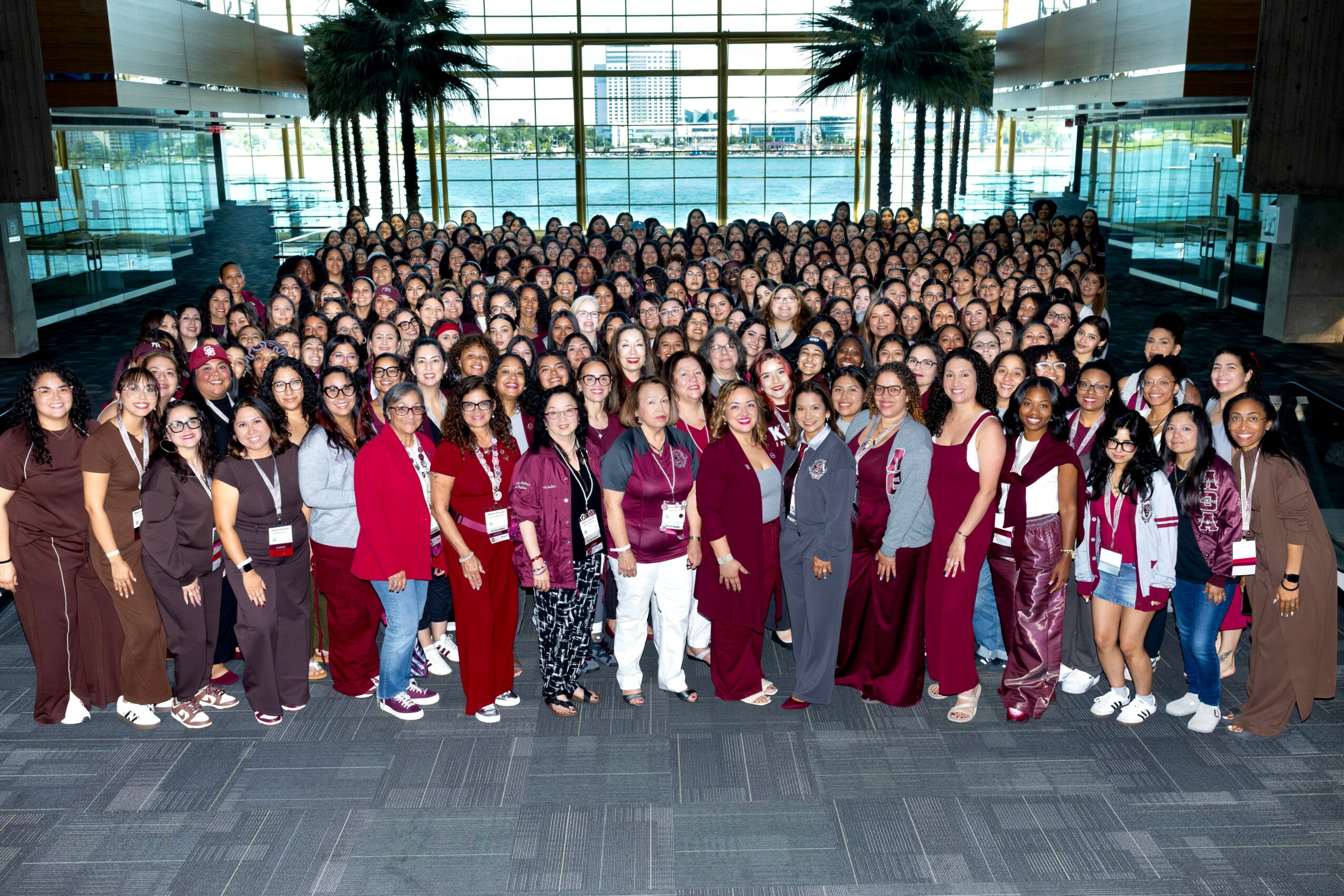 Large group of people gathered indoors in front of glass windows with palm trees outside, posing for a group photo.