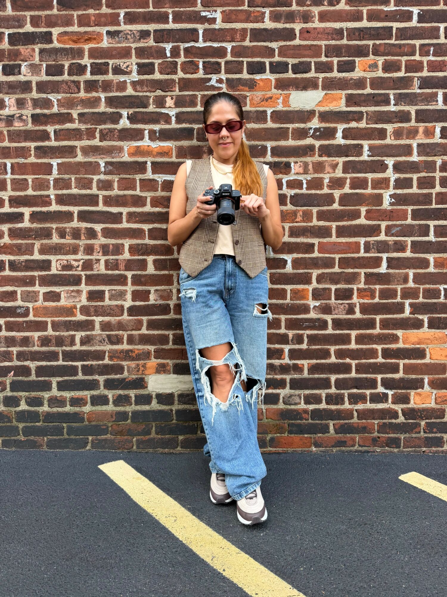 Young woman with sunglasses holding a camera, standing against a brick wall, wearing ripped jeans and sneakers.