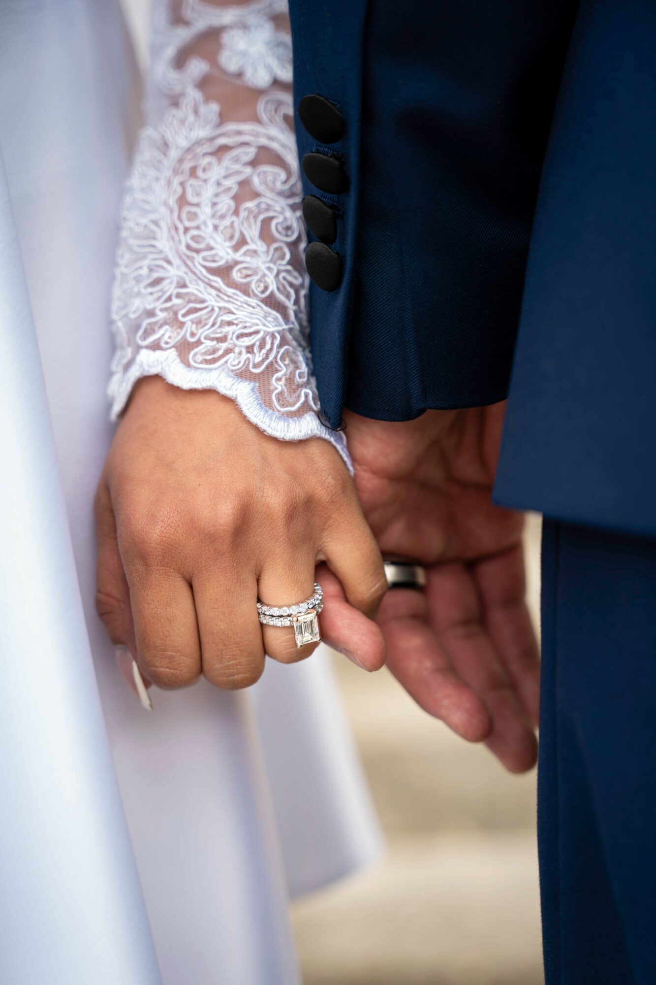 Close-up of a bride and groom holding hands, showing wedding rings and lace sleeve, with the groom in a dark suit.