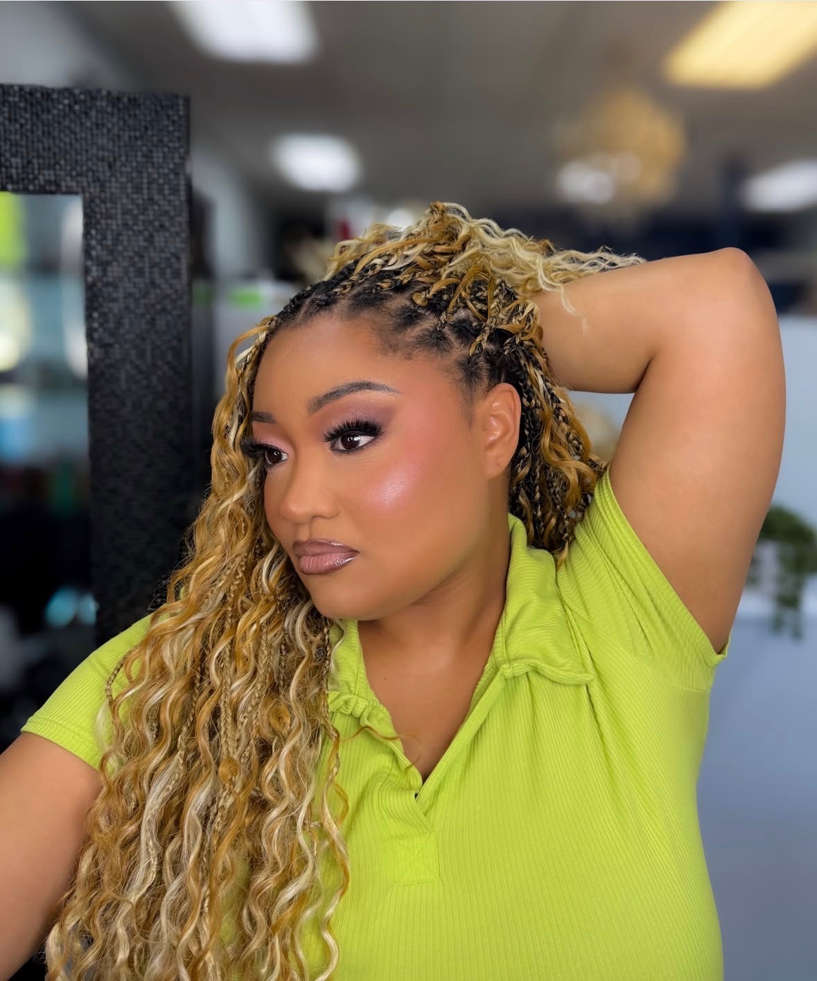 Woman with curly blonde hair and makeup, wearing a bright green shirt, resting her arm on her head indoors.