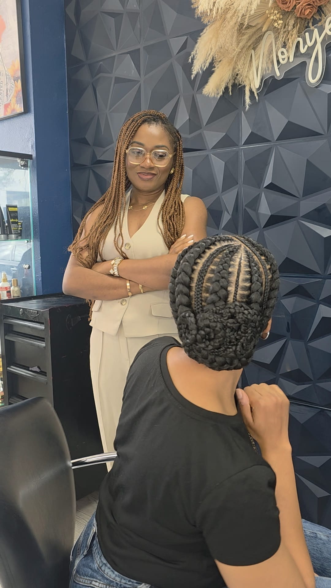 Woman with glasses and long hair standing with arms crossed, facing a person with braided hair in a black shirt, in a modern interior.
