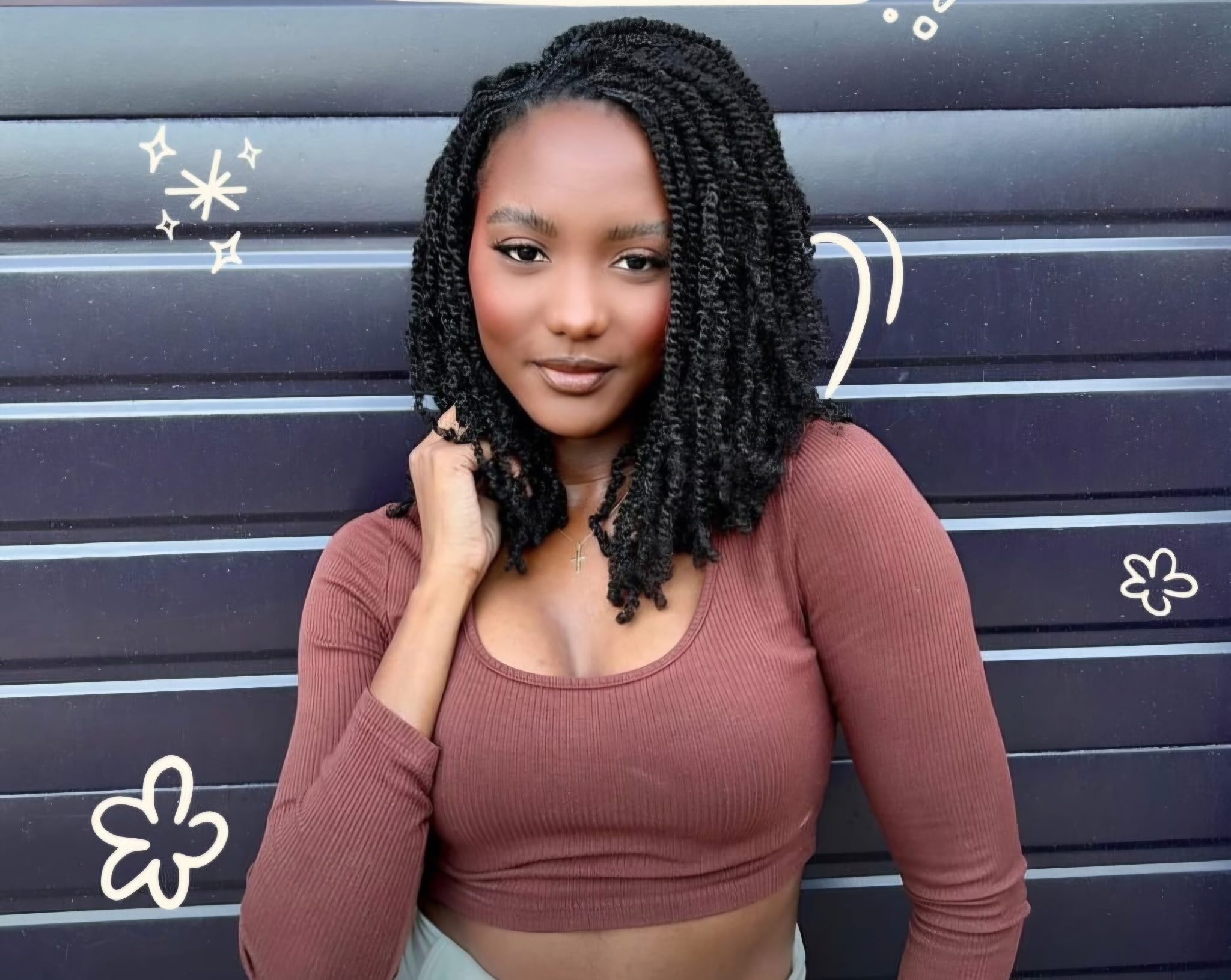 Young woman with shoulder-length curly hair, wearing a brown long-sleeve top, standing in front of a dark wall with white decorative drawings.