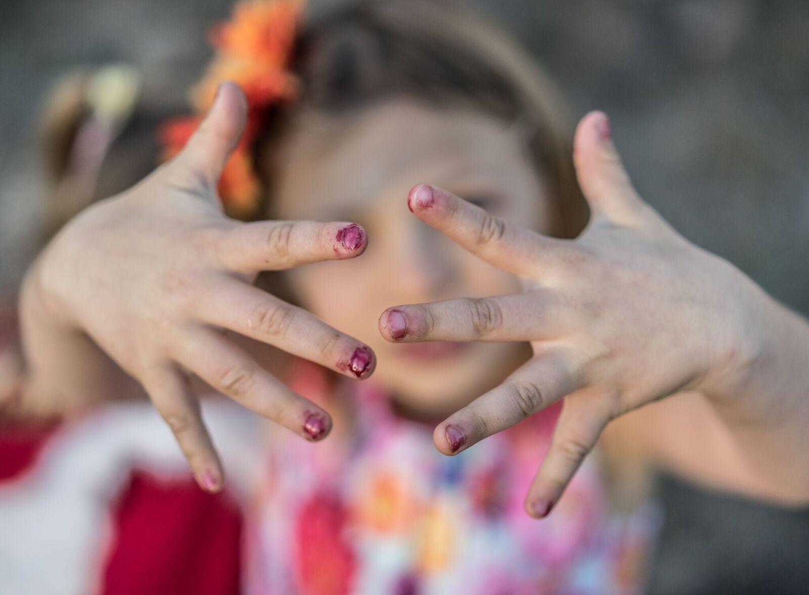 Child with hands covering face, fingers spread, nails painted pink, outdoors, blurred background