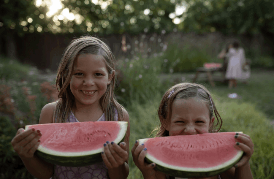 Two children holding watermelon slices outdoors, smiling, with a garden and a person in the background.