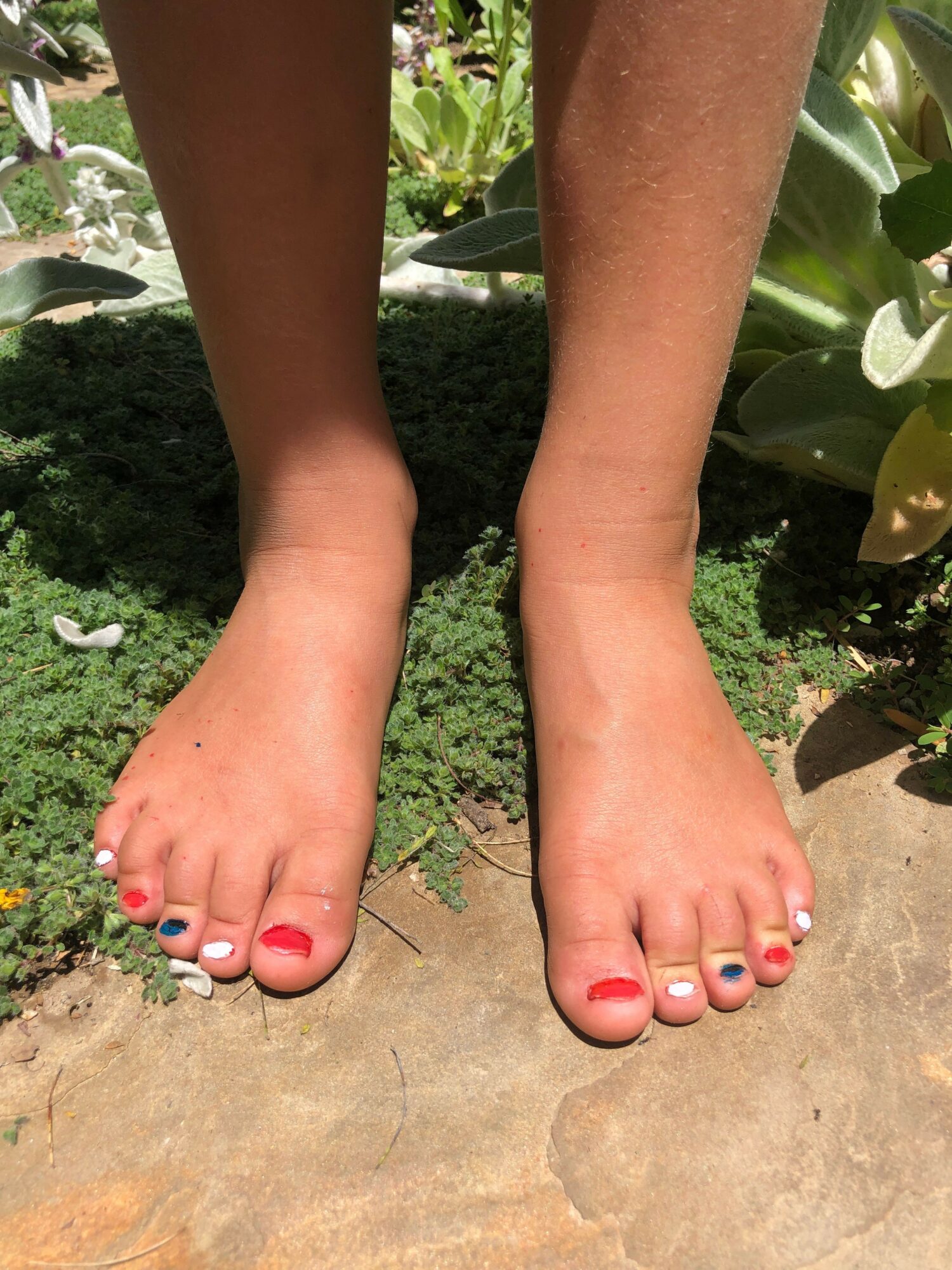 Two feet with painted toenails resting on a stone surface, surrounded by green plants.