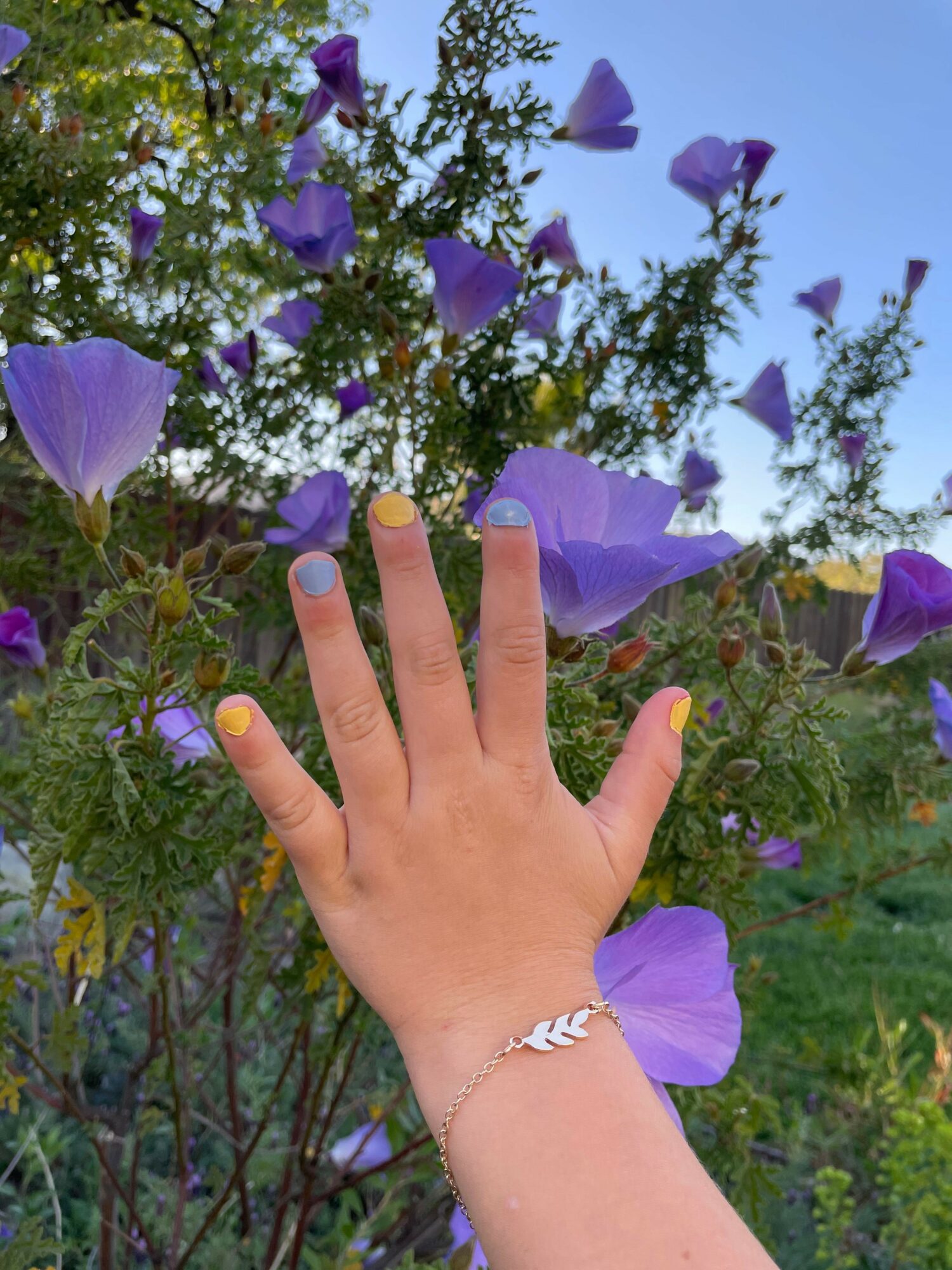 Hand with a bracelet and yellow nail polish reaches toward purple flowers in a garden, blue sky in background.