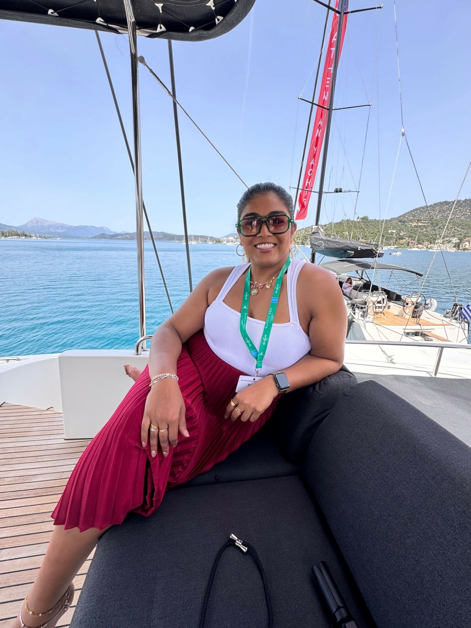Woman in white top and red skirt sitting on a boat with water and boats in background.