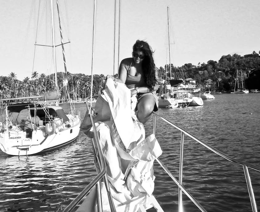 Woman sitting on boat railing holding a dress, with sailboats and trees in the background, black and white photo.
