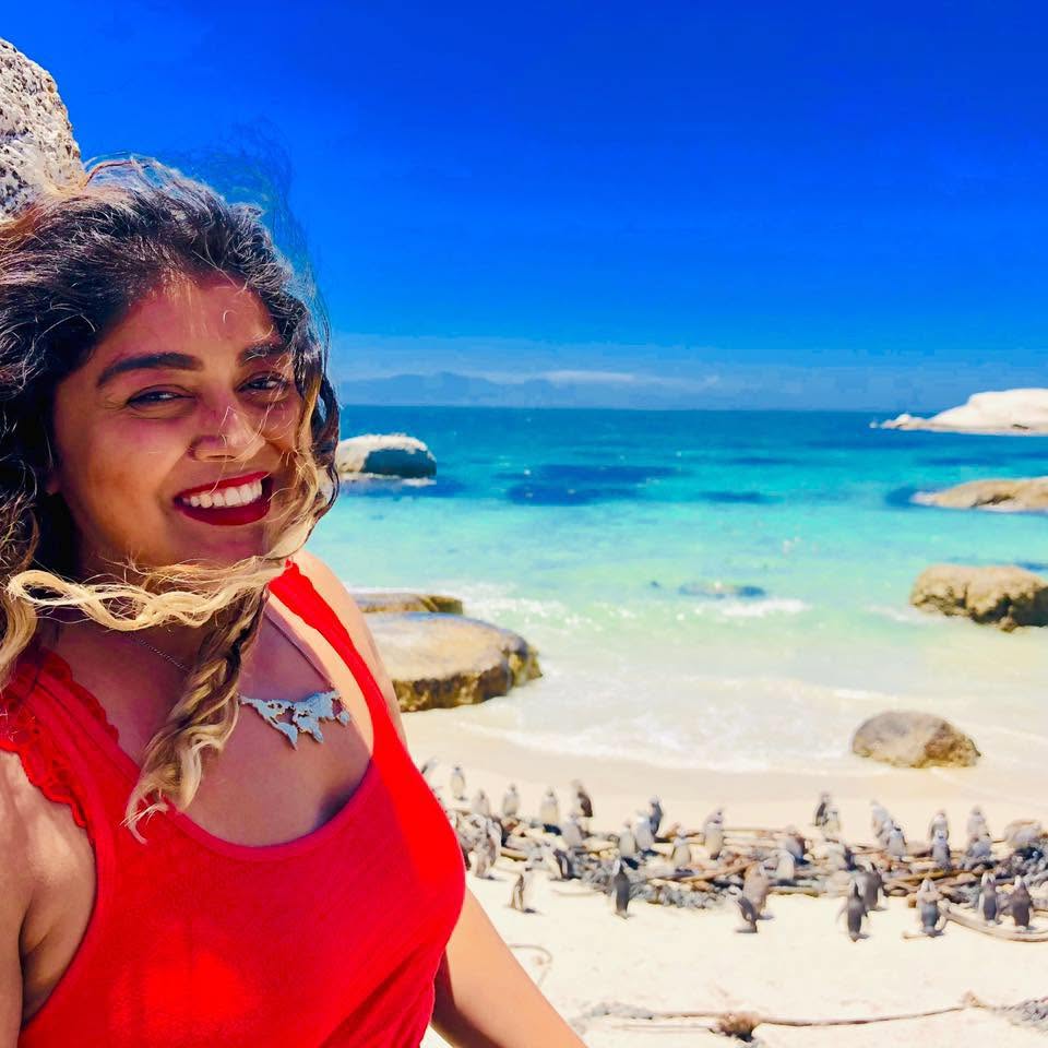 Smiling woman with curly hair in a red top at the beach, ocean and rocks in background.
