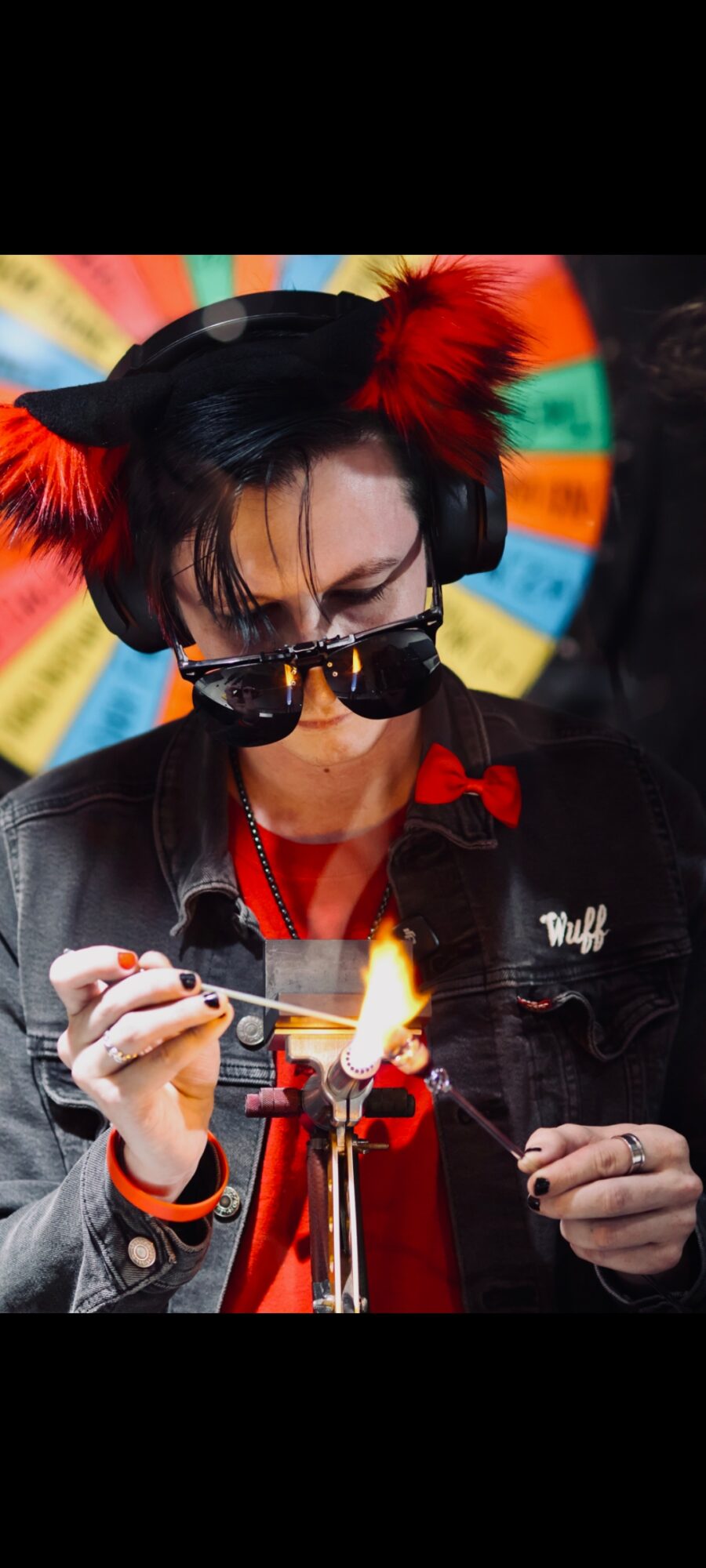 Person with black hair, wearing cat ears with red feathers, sunglasses, and a black jacket, lighting a firework.