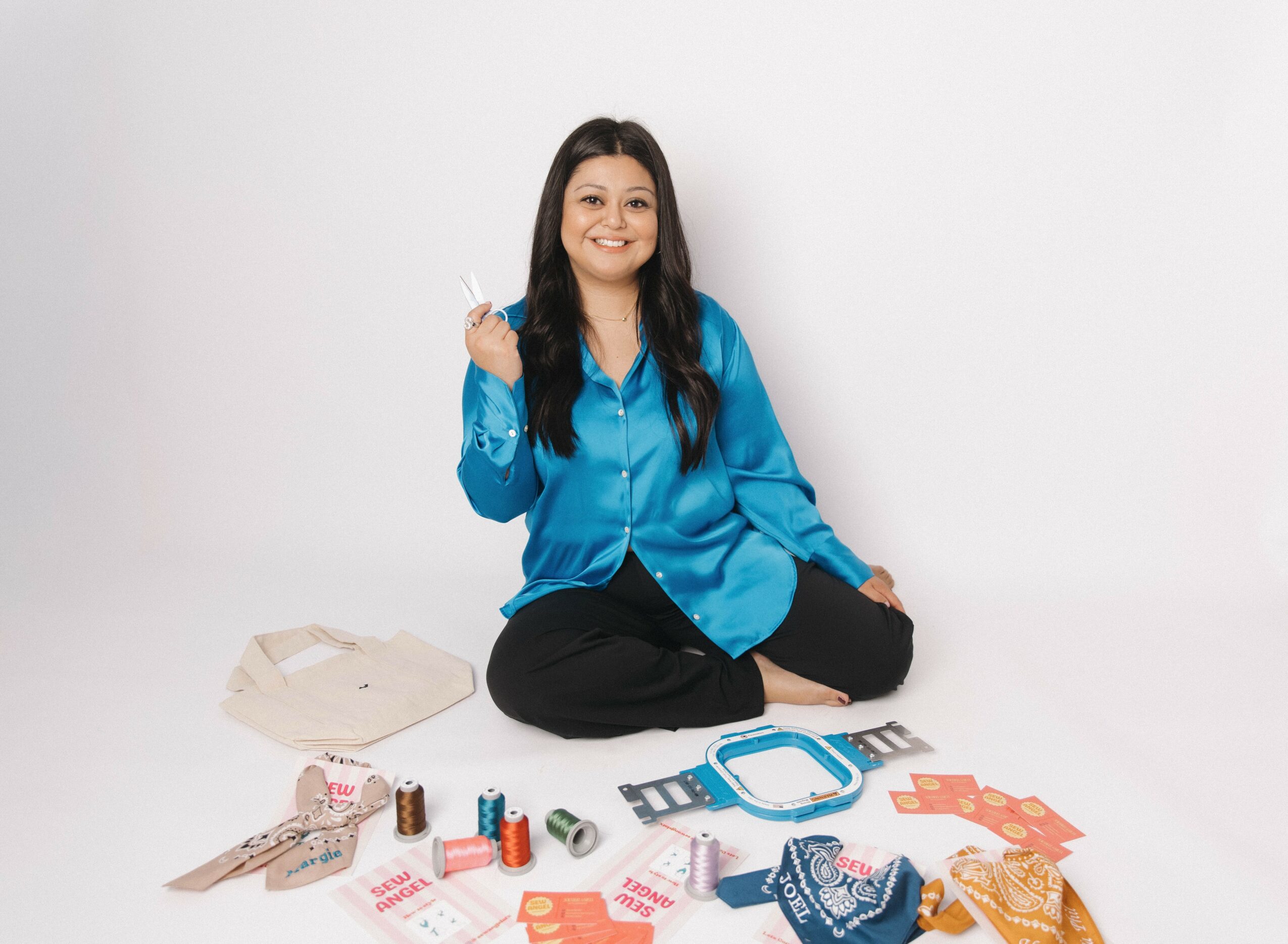 Woman sitting cross-legged on the floor with various makeup and beauty products spread around her, smiling, against a plain white background.
