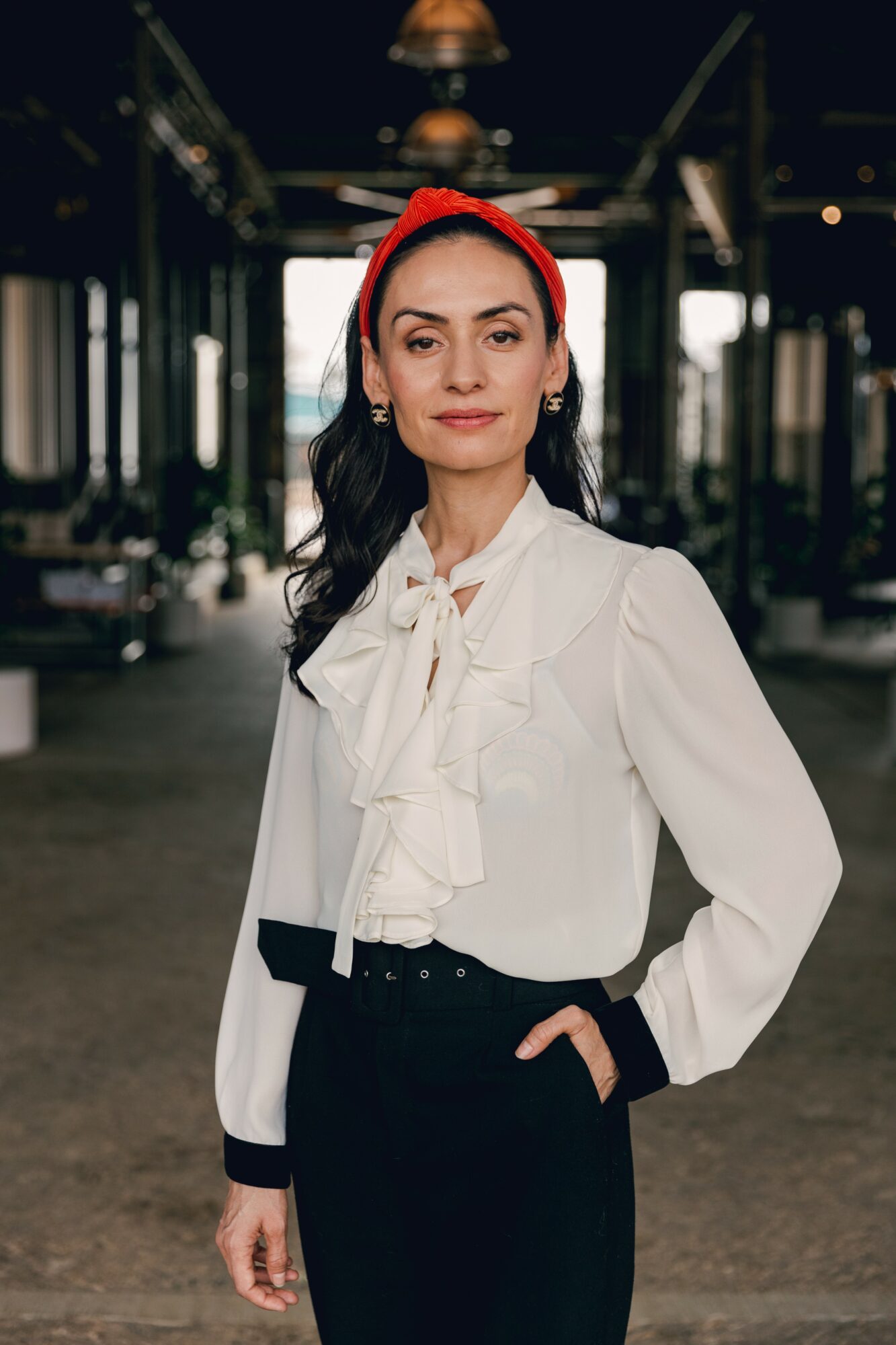 Woman with dark hair, red headband, white blouse with bow, black pants, standing indoors with industrial background.
