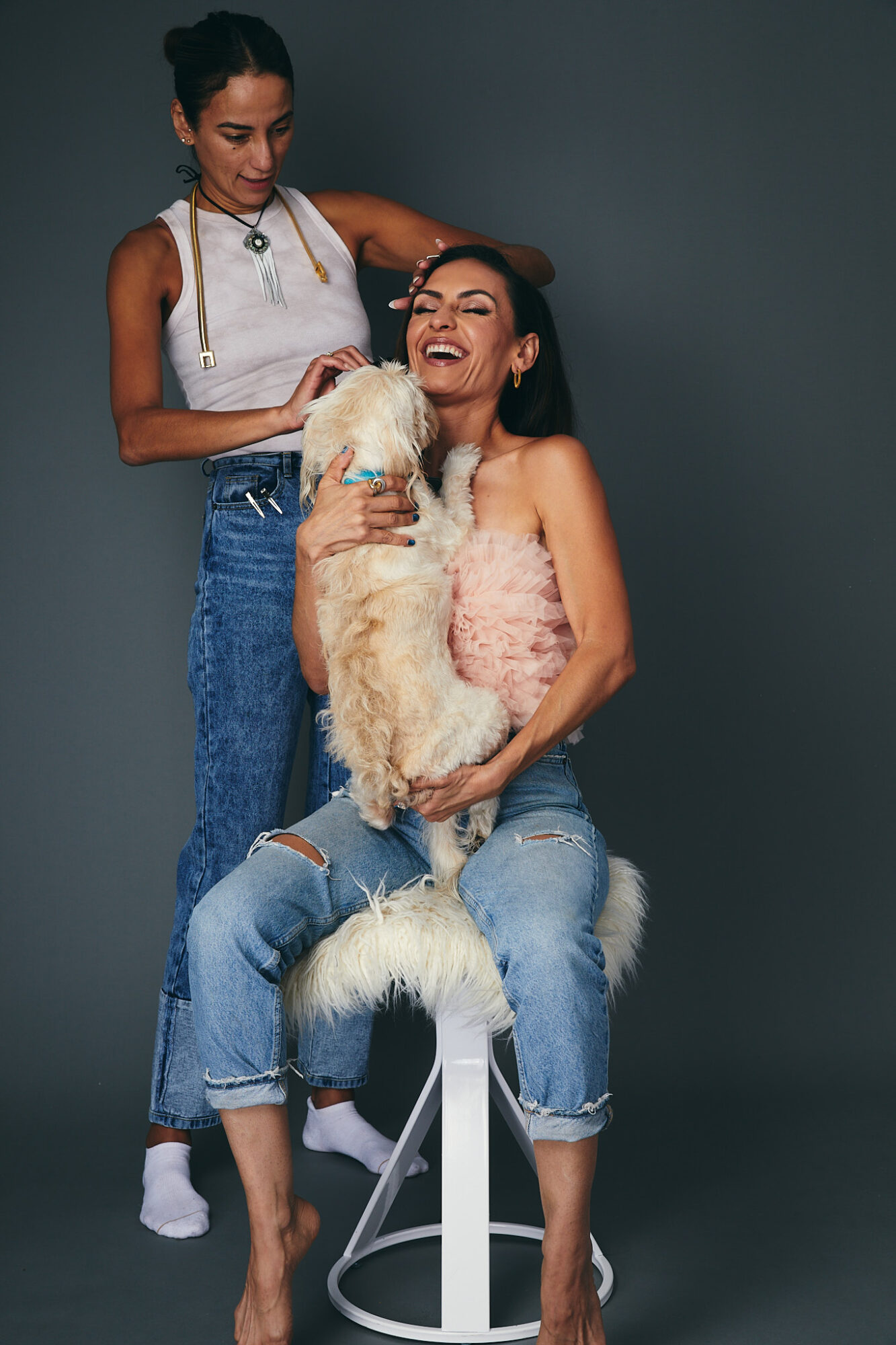 Two women and a dog, one woman sitting on a stool, the other standing, with a dark background.