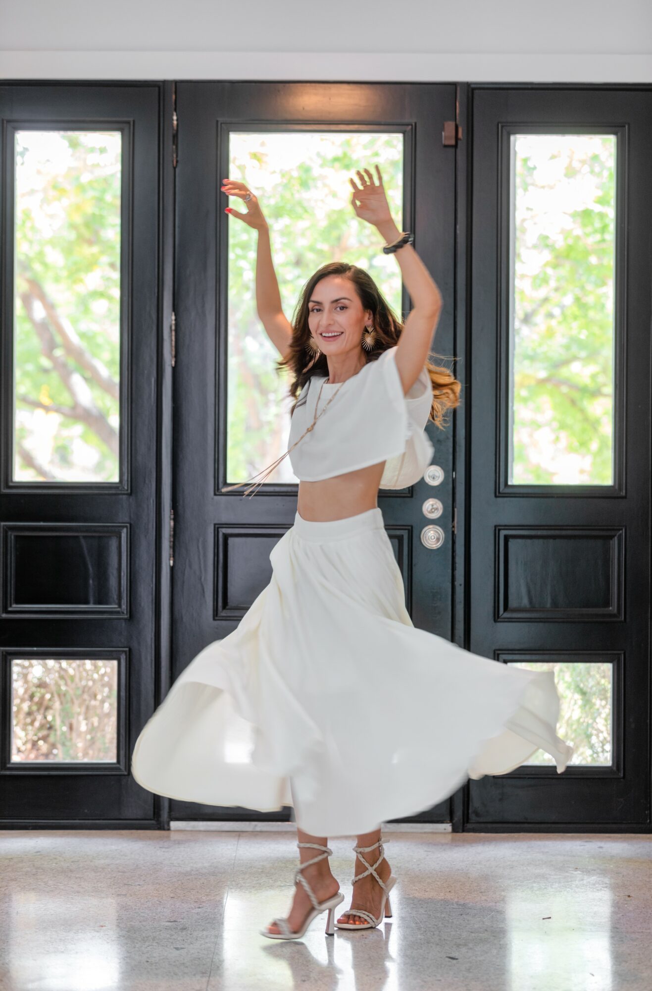 Woman dancing indoors with arms raised, wearing a white skirt and top, in front of large windows with trees outside.