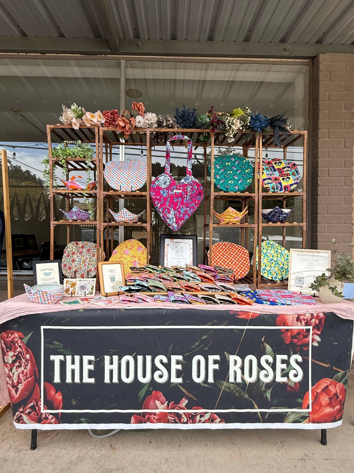 Table with a banner reading 'The House of Roses' displays colorful items, with shelves of flowers and decorations behind it.