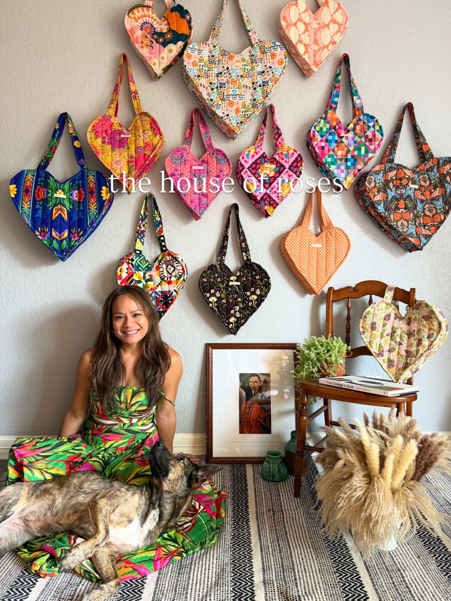 Woman sitting on floor with two dogs, colorful bags on wall, framed photo, plant, and decorative items in room.