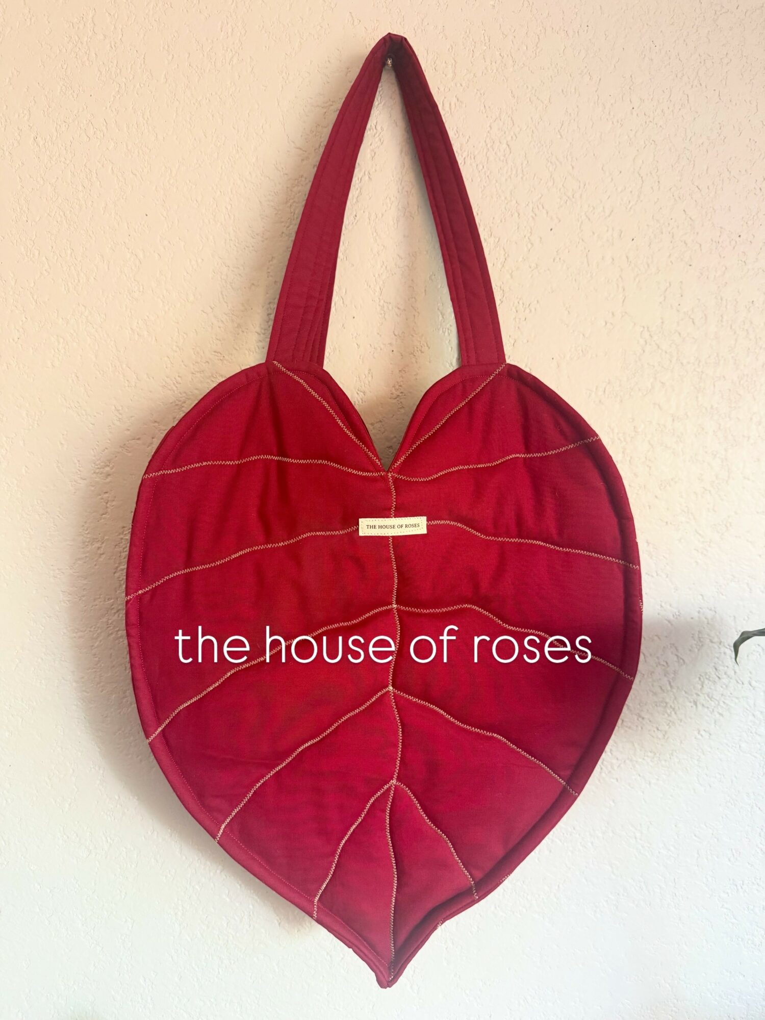 Red heart-shaped bag with white stitching and text reading 'the house of roses' on a plain wall.