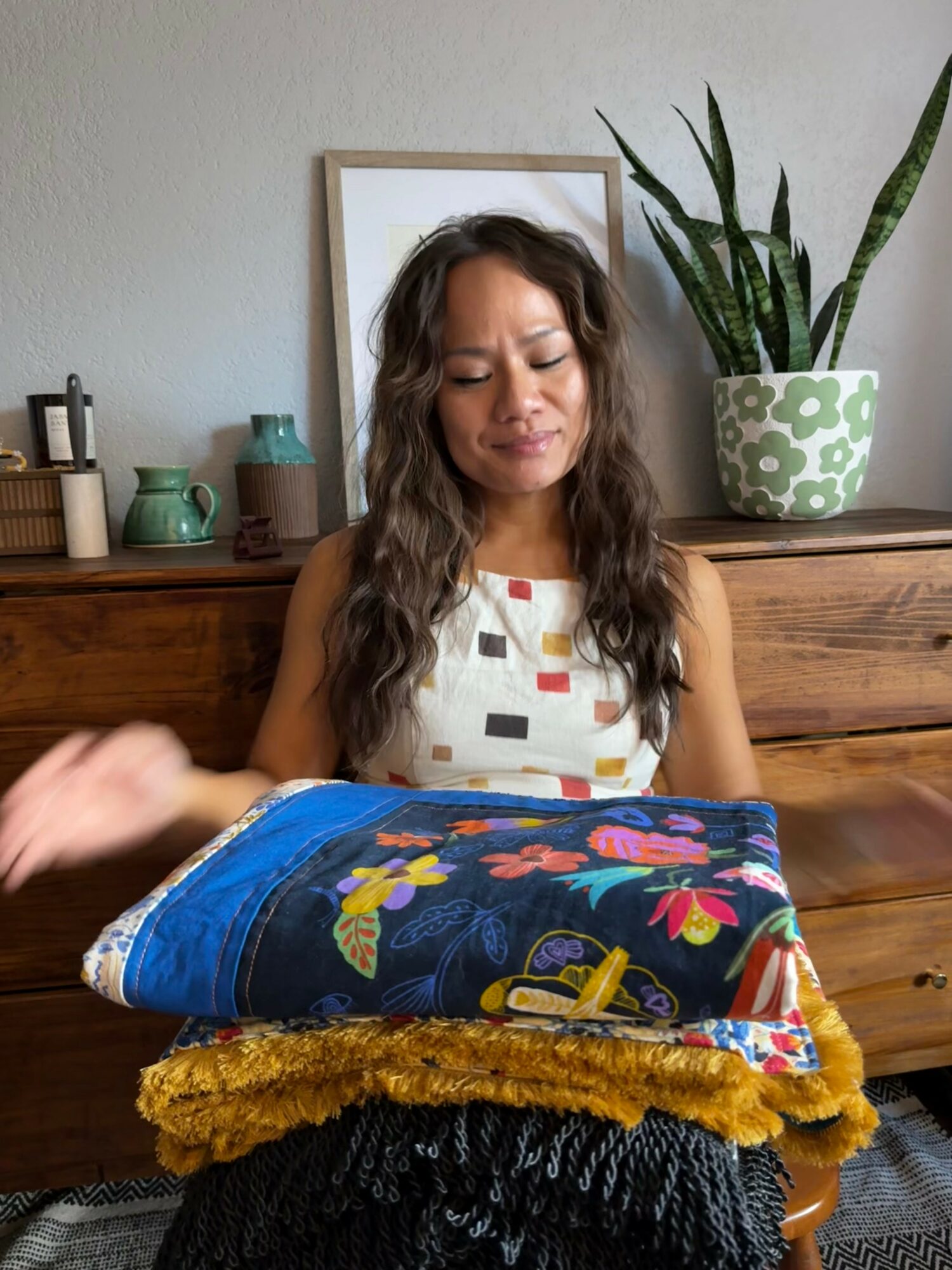 Woman with long hair sitting at a table with folded colorful blankets, in a room with a plant and framed picture behind.