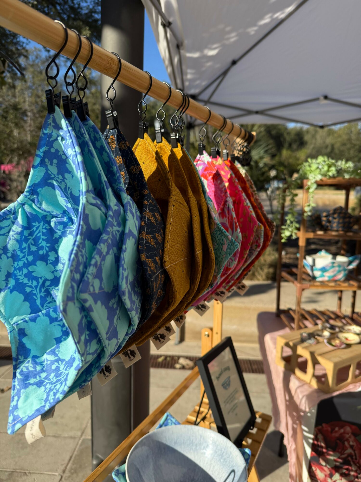 Colorful fabric face masks hanging on a bamboo rack outdoors, with other items and furniture in the background.