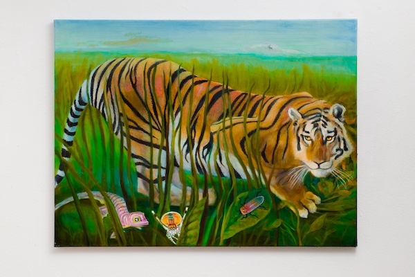 A tiger lying in tall grass with a landscape background, including a blue sky and distant water, and objects in the grass.