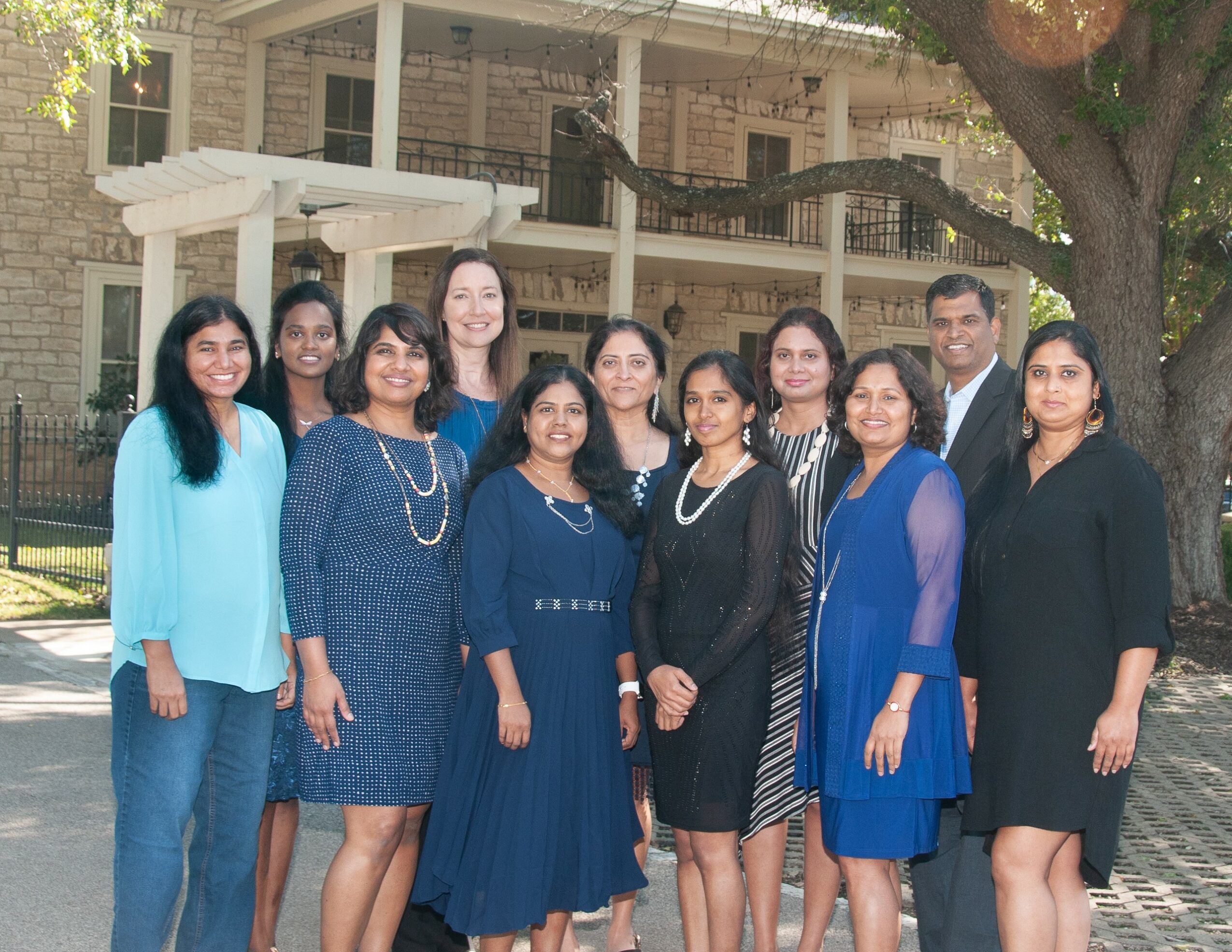 Group of ten women and one man standing outdoors in front of a building and tree.