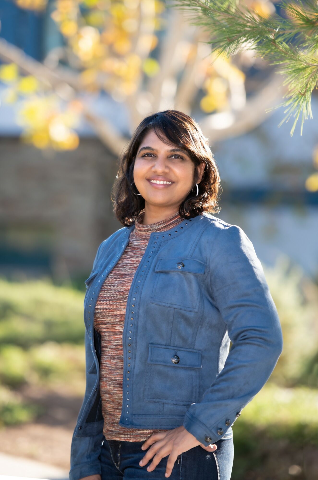 Woman smiling outdoors with trees and sunlight in background, wearing a denim jacket and striped shirt.