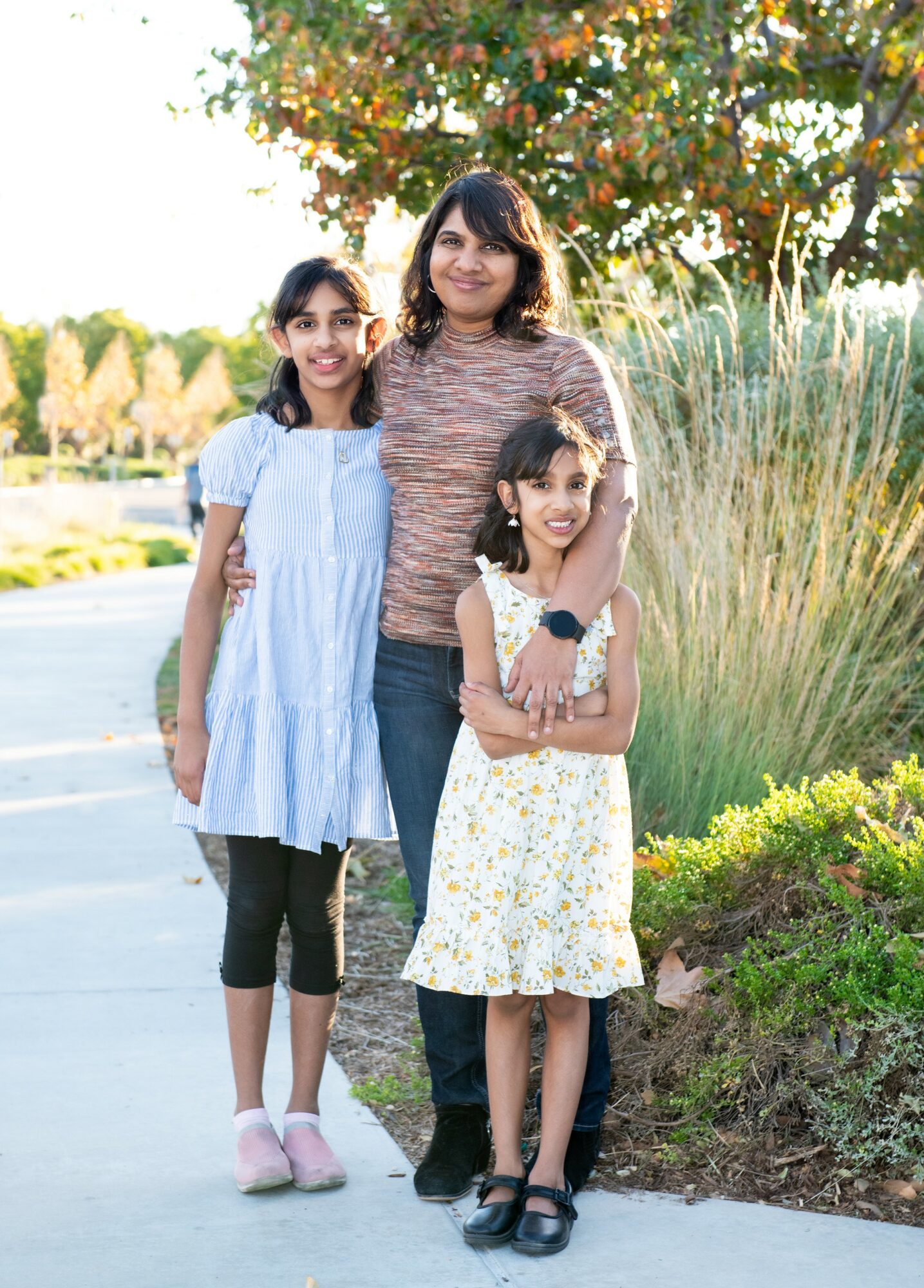 Three people, a woman and two girls, standing outdoors on a sidewalk with trees and plants in the background.