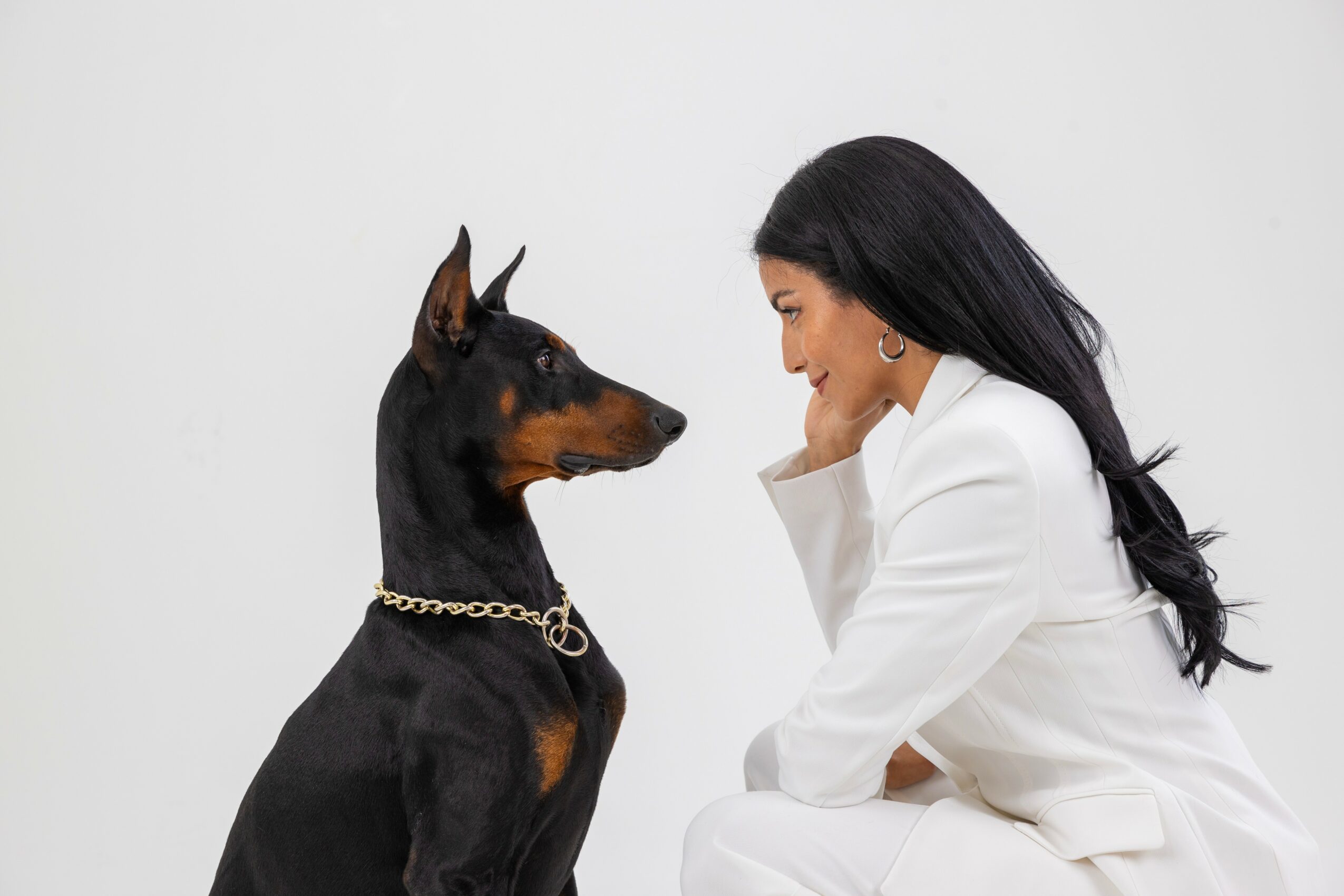 Woman with long black hair smiling at a Doberman dog wearing a chain collar, both facing each other.