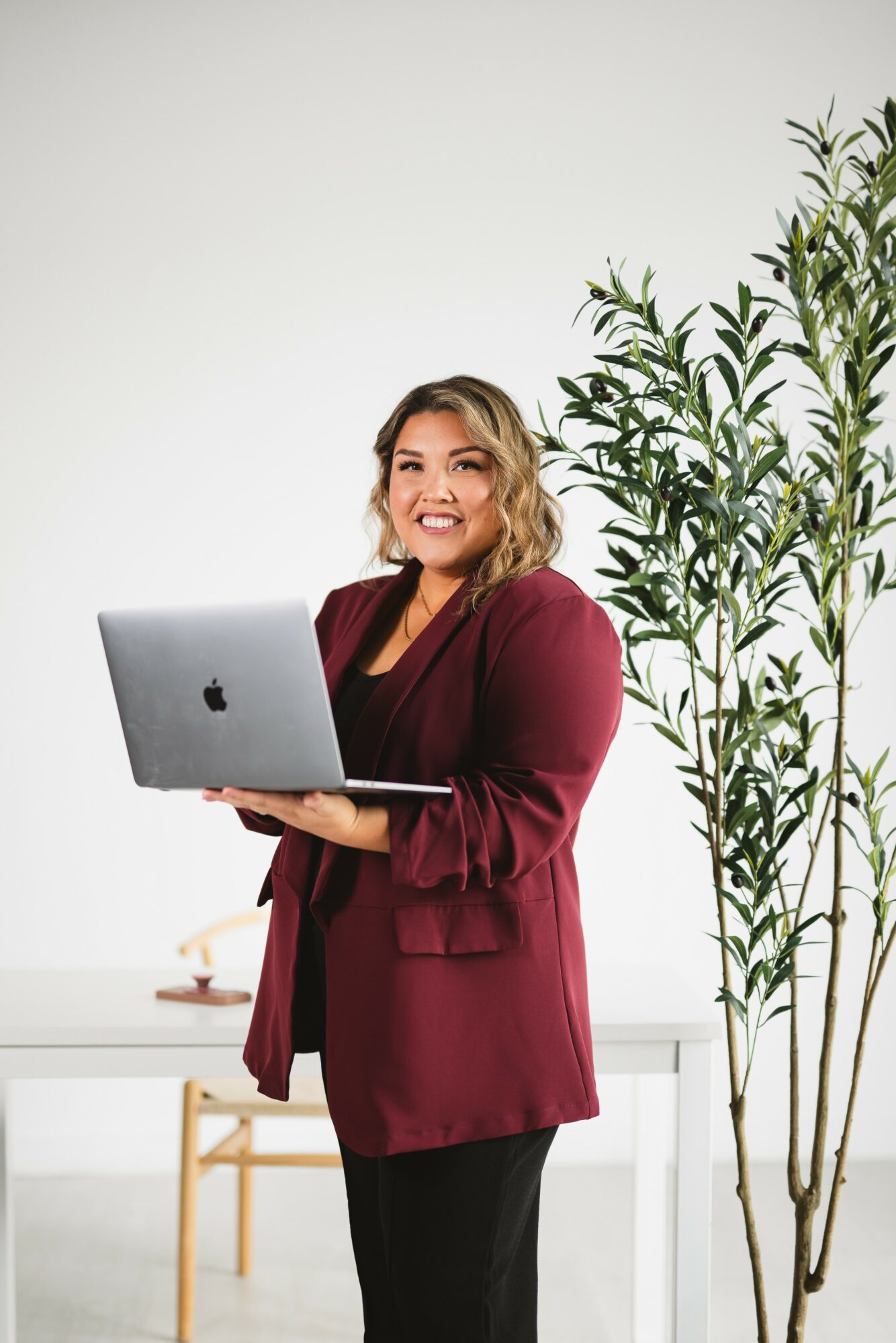 Woman smiling holding a laptop, standing next to a tall plant in a bright room.