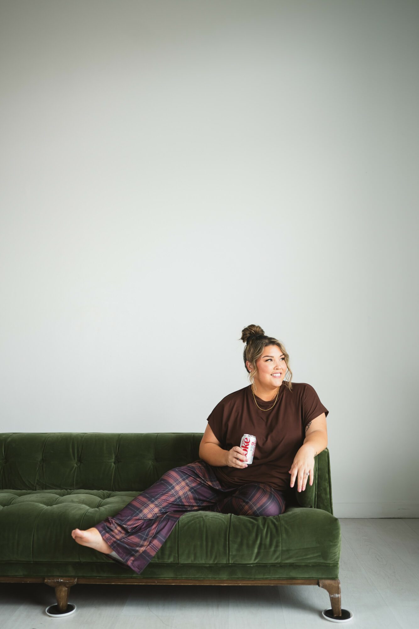 Woman sitting on a green sofa holding a can, smiling, against a plain light-colored wall.