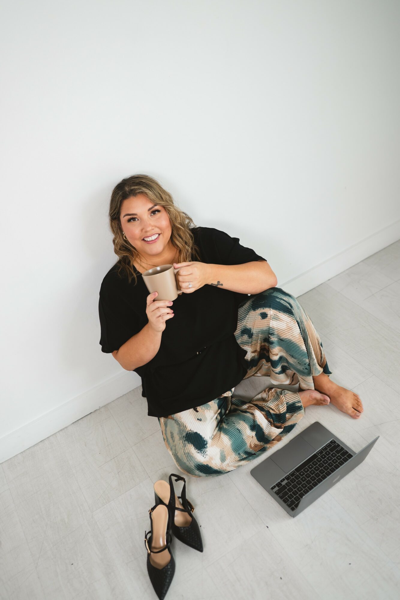 Person sitting on floor against white wall, holding a mug, with a laptop nearby, smiling, wearing black top and patterned pants.