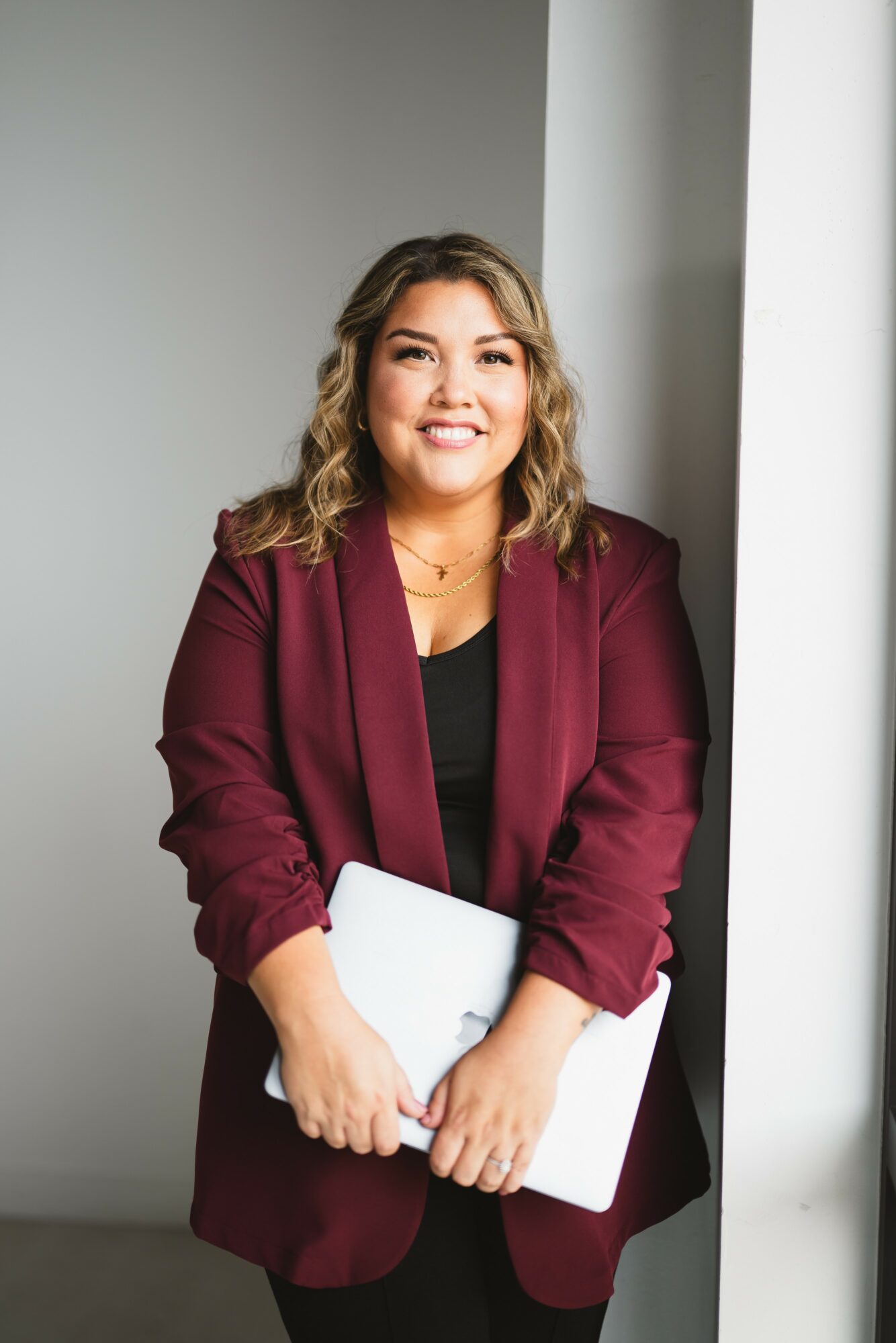 Smiling woman with wavy hair holding a laptop, wearing a maroon blazer and black top, standing near a white column.
