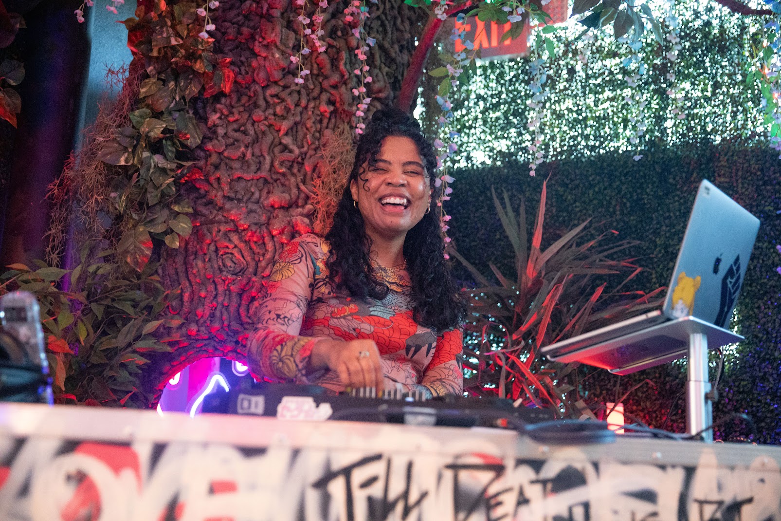 Woman with curly hair smiling behind a DJ setup outdoors with trees and plants in background.