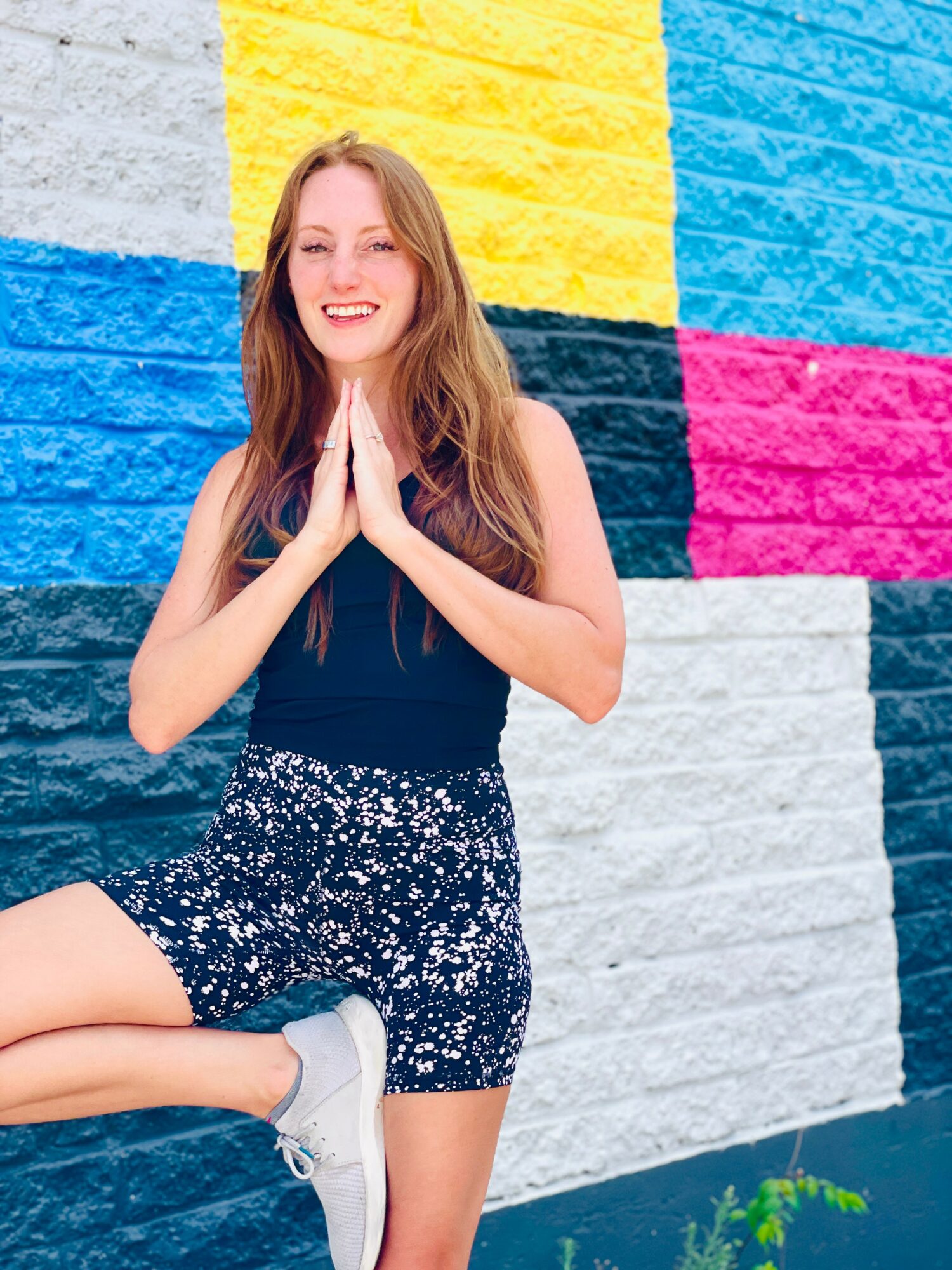 Young woman with long hair smiling, hands together, standing in front of a colorful brick wall.