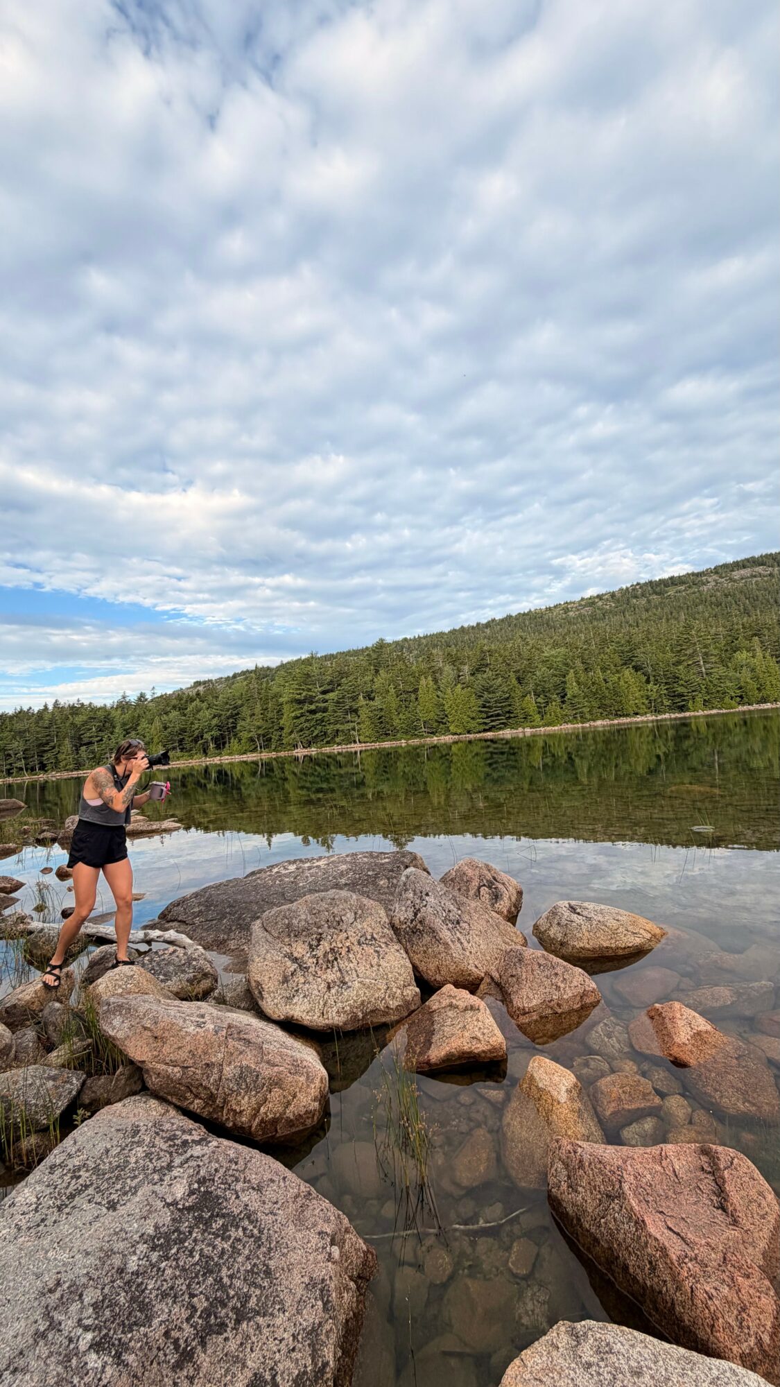 Person standing on rocks by a calm lake with a forested hill and cloudy sky in the background.