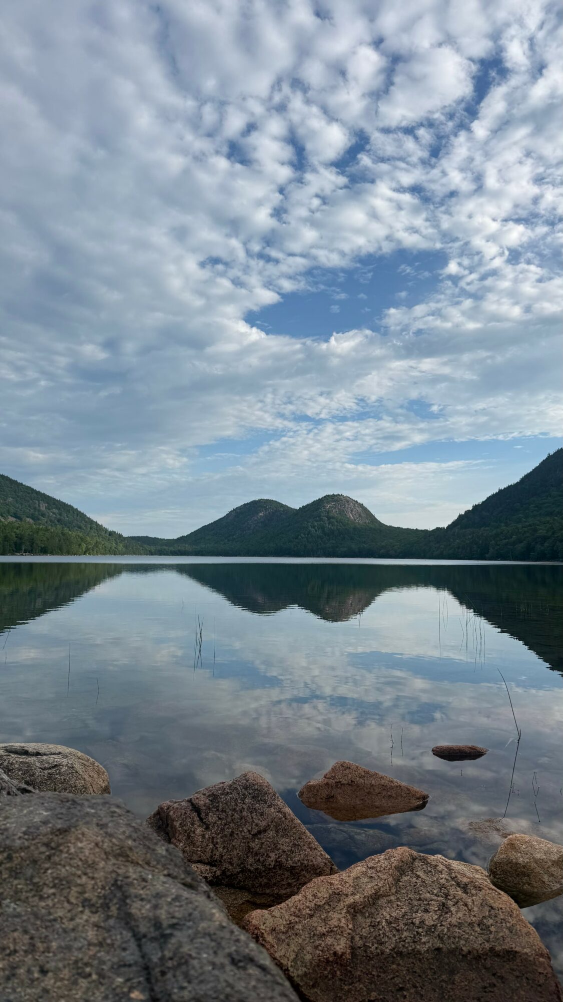 Calm lake reflecting mountains and partly cloudy sky, with rocks in the foreground.