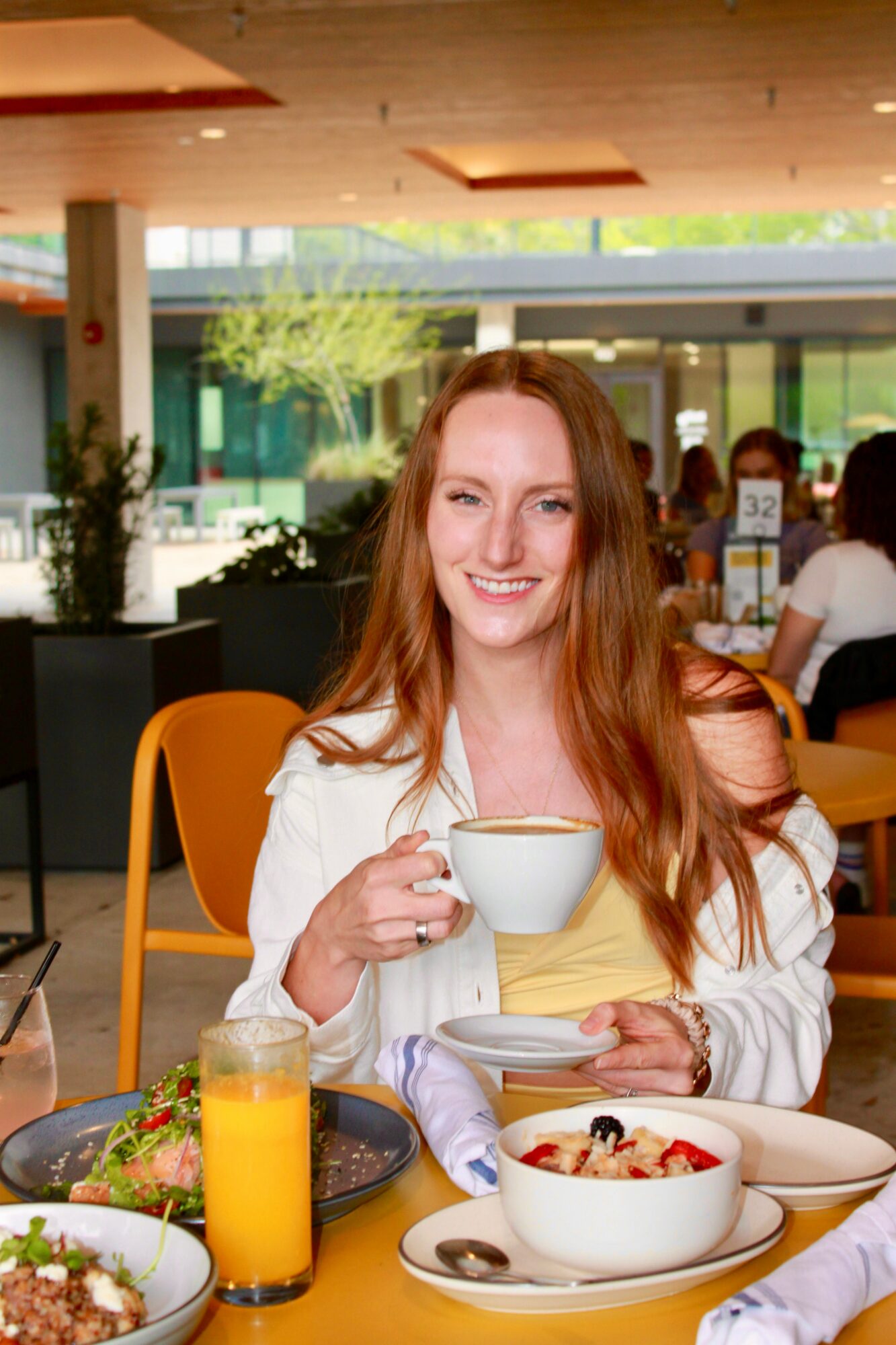 Young woman with long red hair smiling, holding a cup, sitting at a table with breakfast foods in a cafe.
