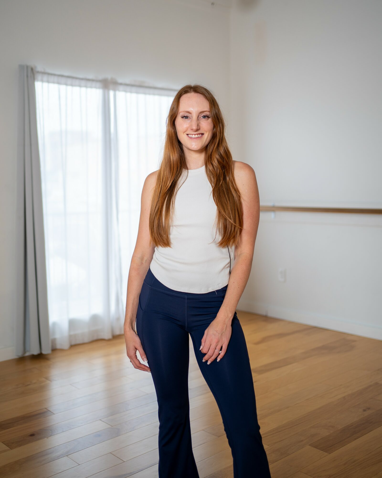Smiling woman with long hair standing in a bright room with wooden floor and large window, wearing a white top and dark pants.