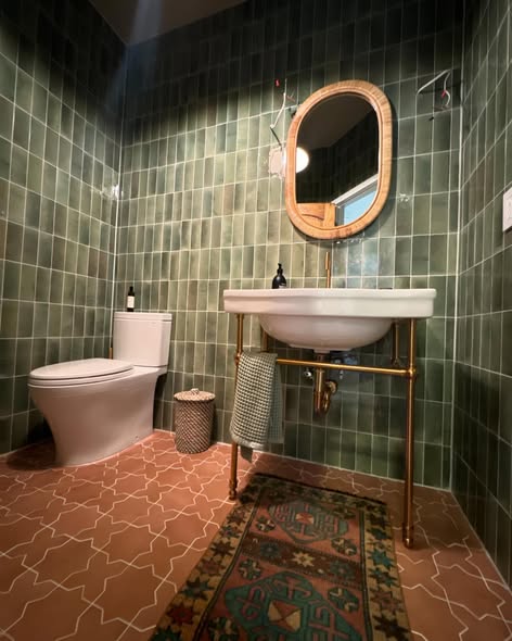 Bathroom with green tiled walls, toilet, sink with mirror, small basket, and patterned rug.