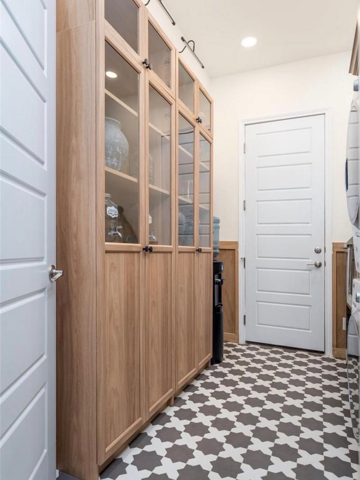 Tall wooden cabinet with glass doors in a room with patterned floor and white door at the end.