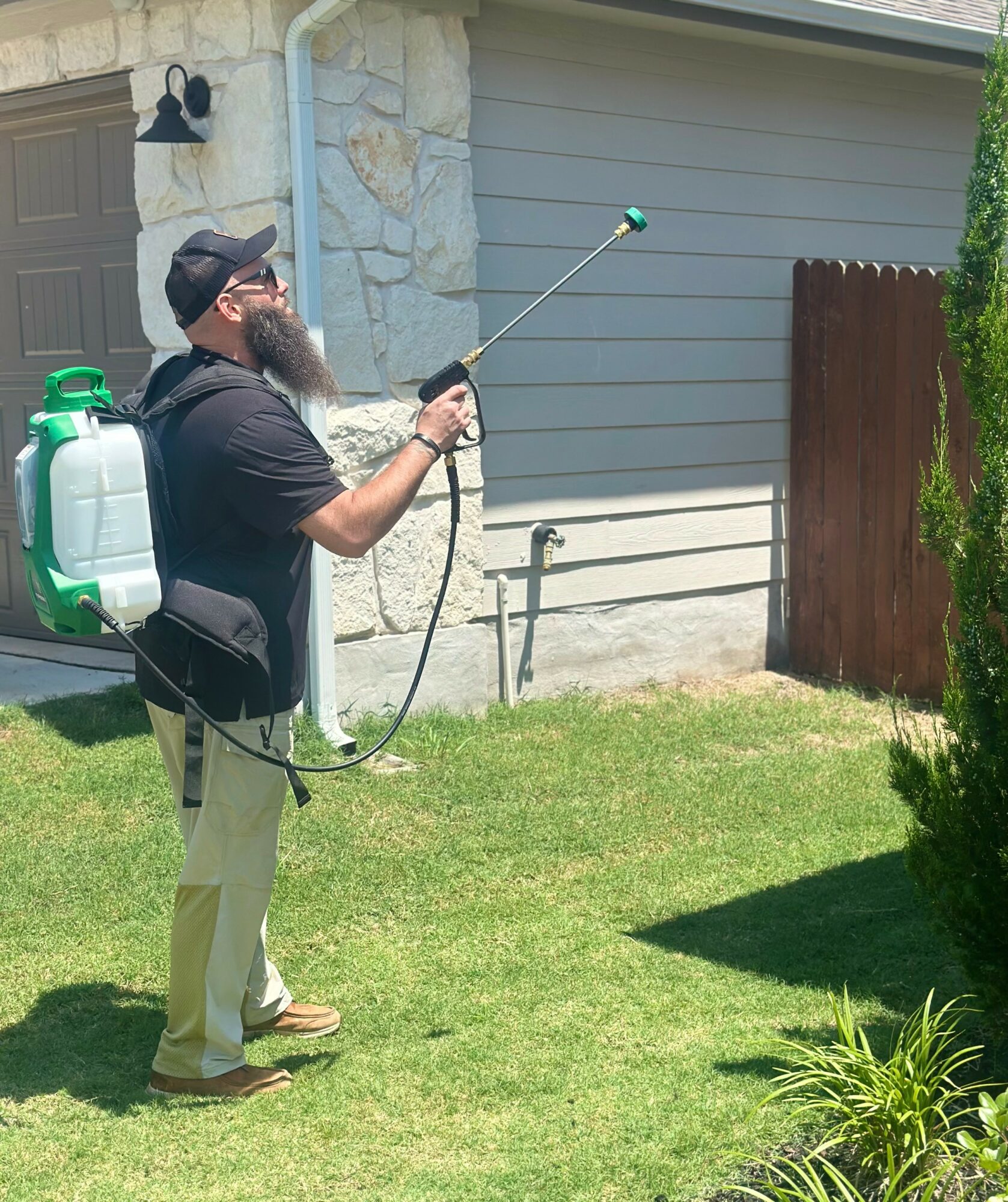 Man with a beard wearing a cap and black shirt sprays a house exterior with a garden sprayer, standing on grass.