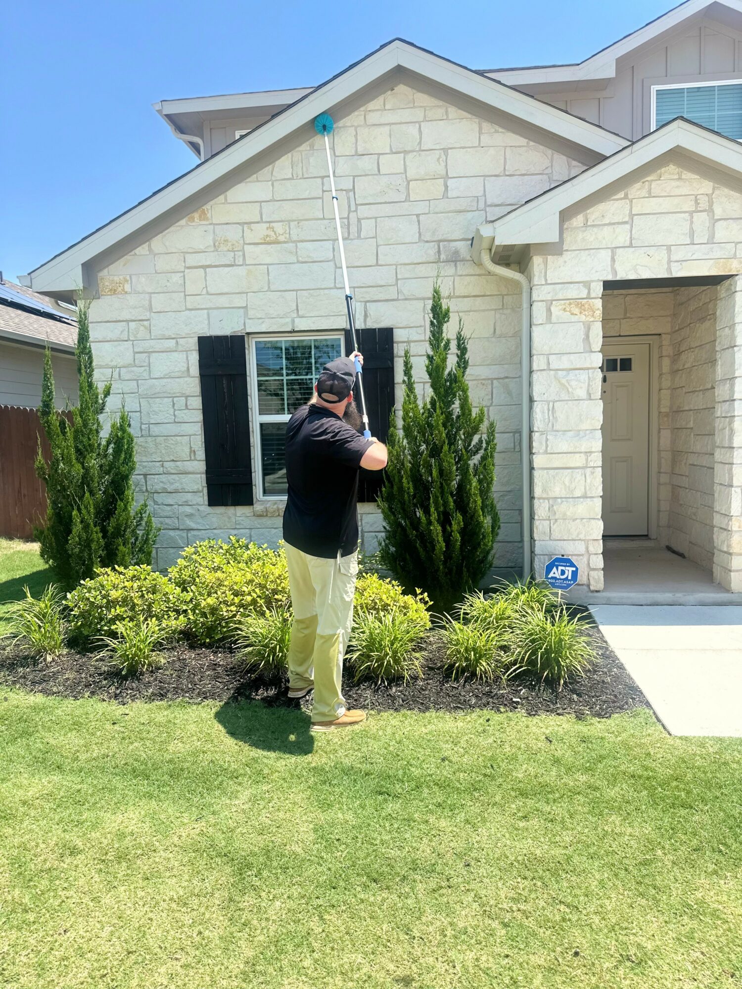 Man standing in front of a house with a garden, holding a long pole, wearing a hat and sunglasses.
