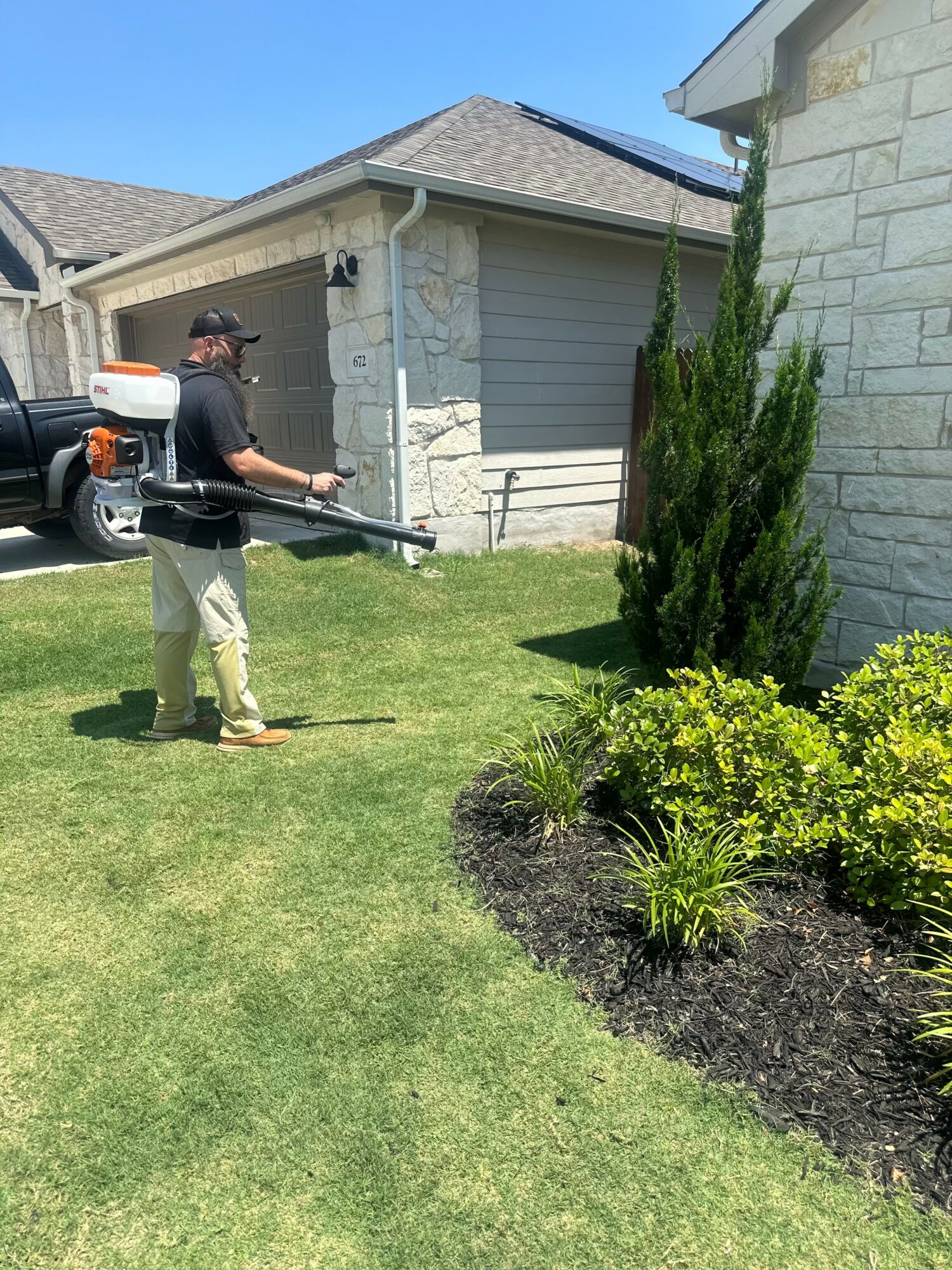 Person using a leaf blower on a lawn next to a house with a stone exterior and a small garden bed with plants.