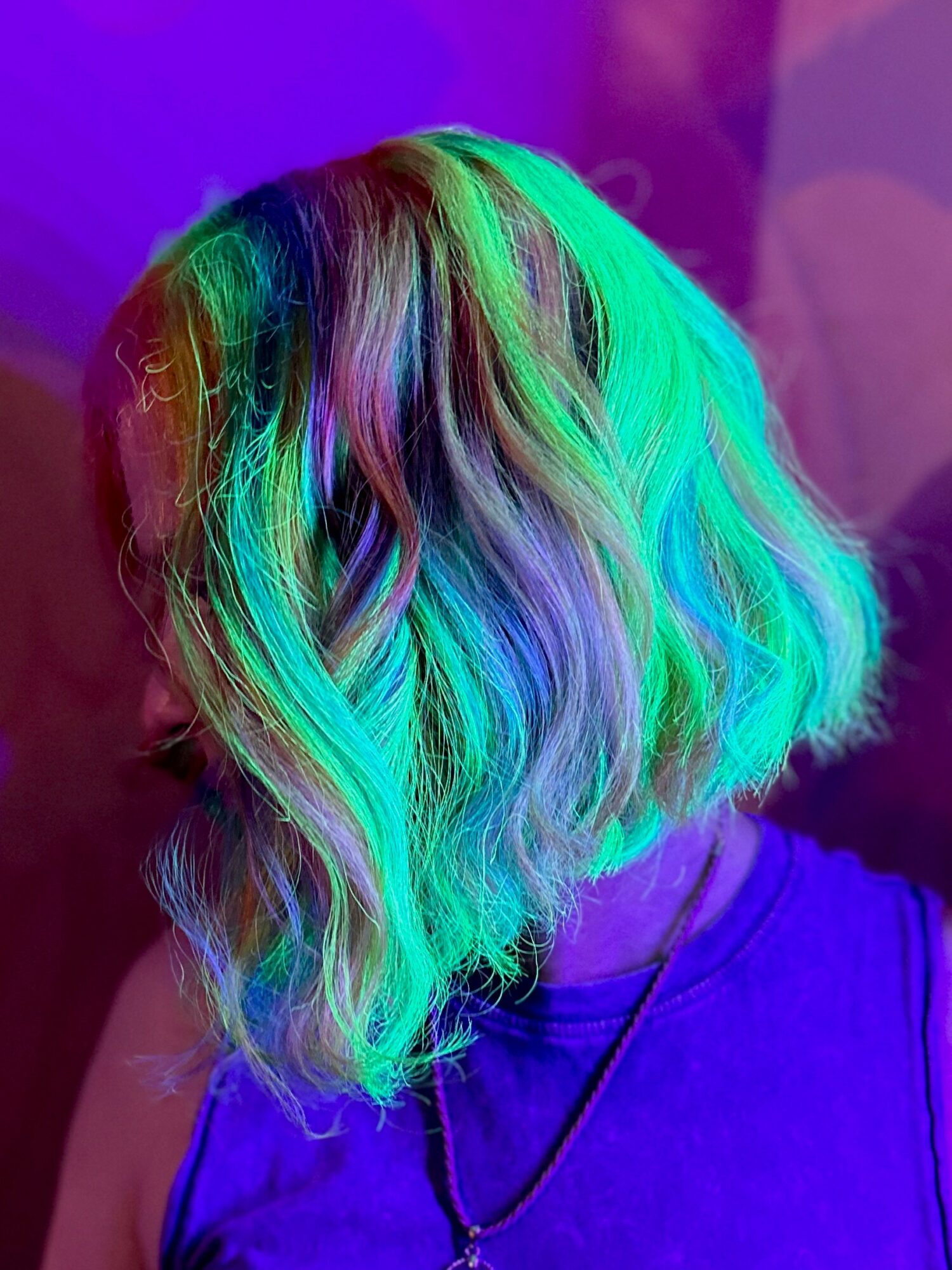 Colorful wavy hair with shades of green, purple, and blue, illuminated by vibrant lighting, worn by a person in a purple top.