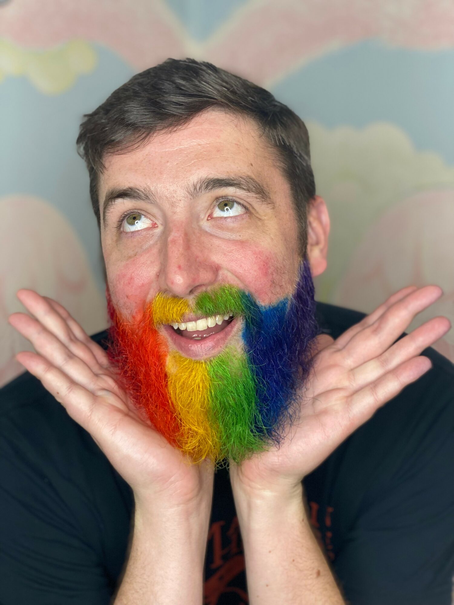 Person with rainbow-colored beard and makeup, smiling with hands near face, colorful background.