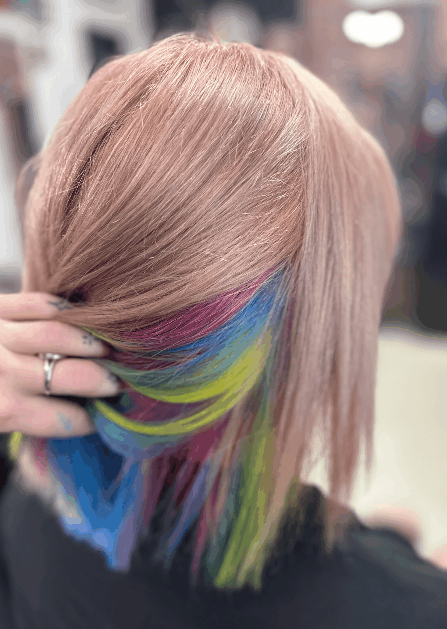 Person with pastel pink hair and rainbow-colored tips, holding a section of hair, in a salon setting.